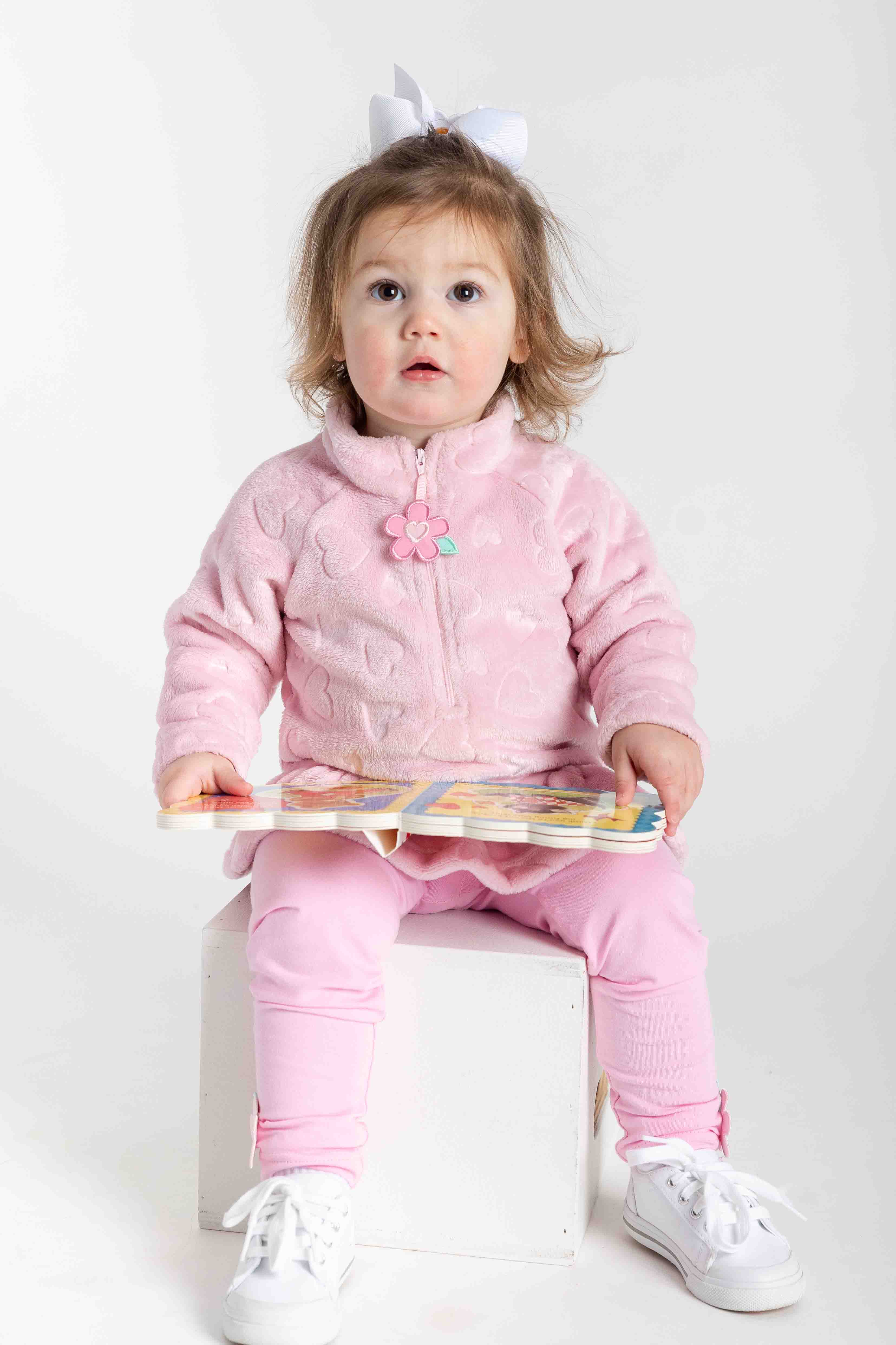 Child wearing a pink outfit with a bow, sitting on a white block holding a book.