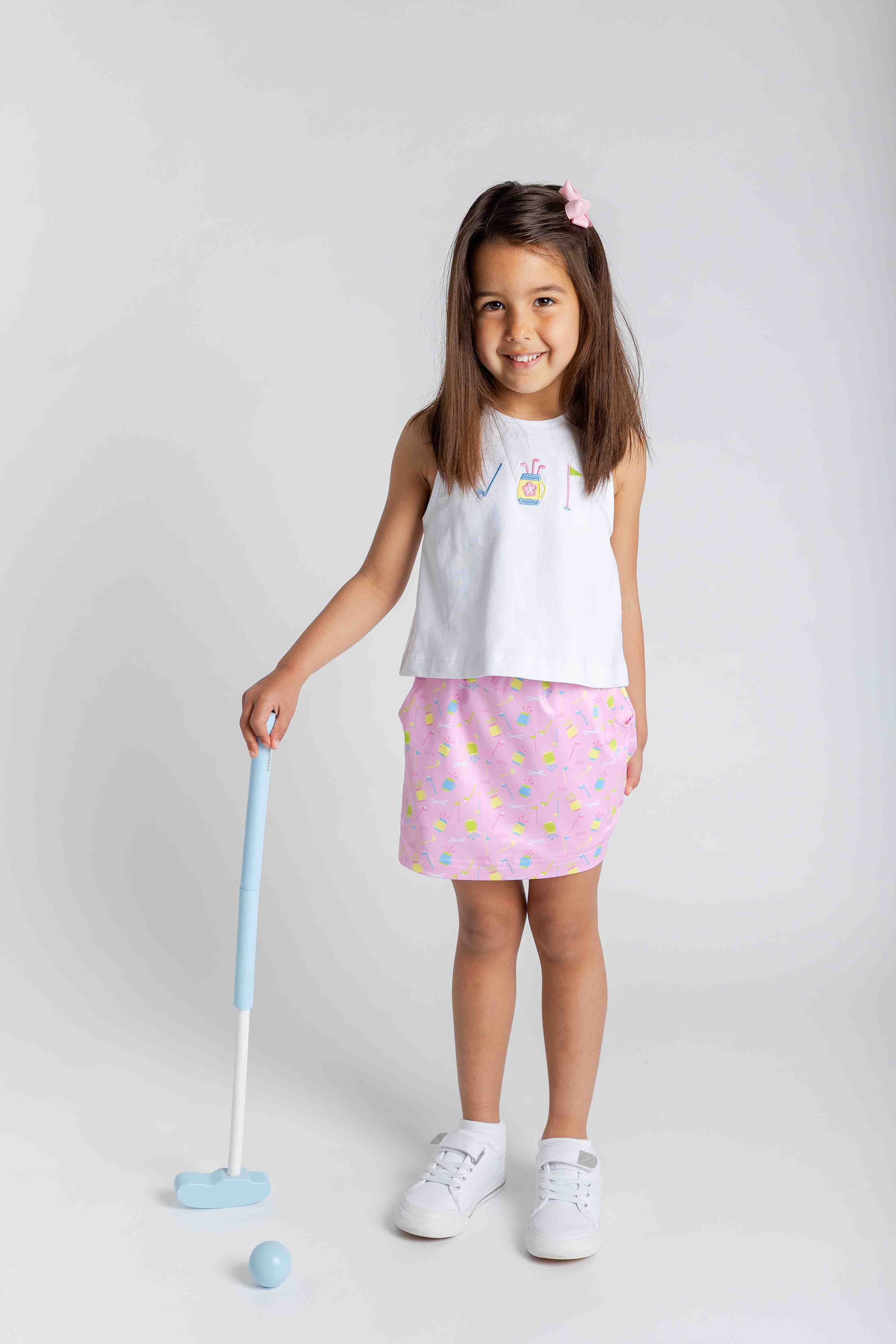 Young girl in a white sleeveless top and pink skirt holding a golf club on a plain background
