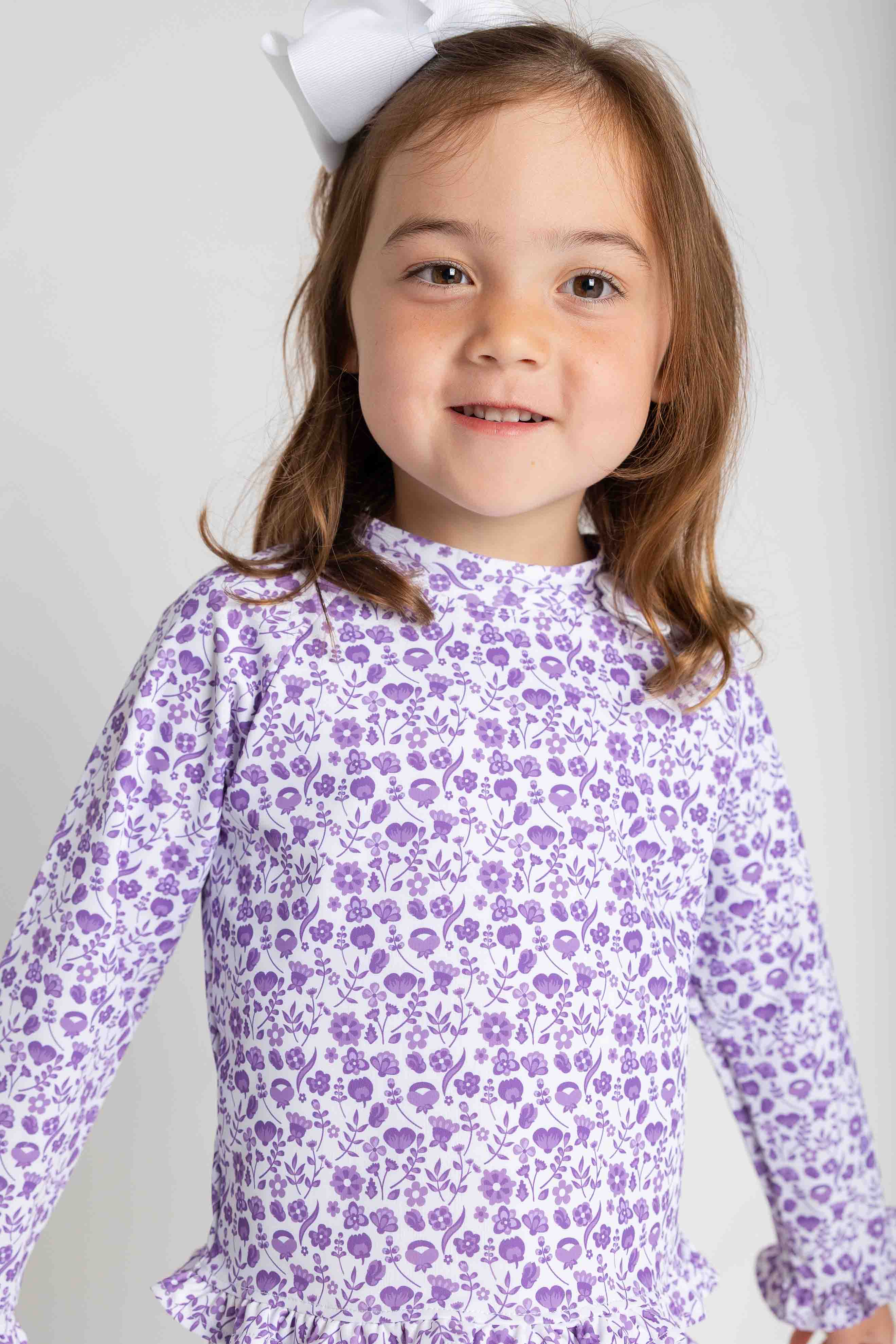 Young girl wearing a purple floral dress with a white bow on her head against a plain background