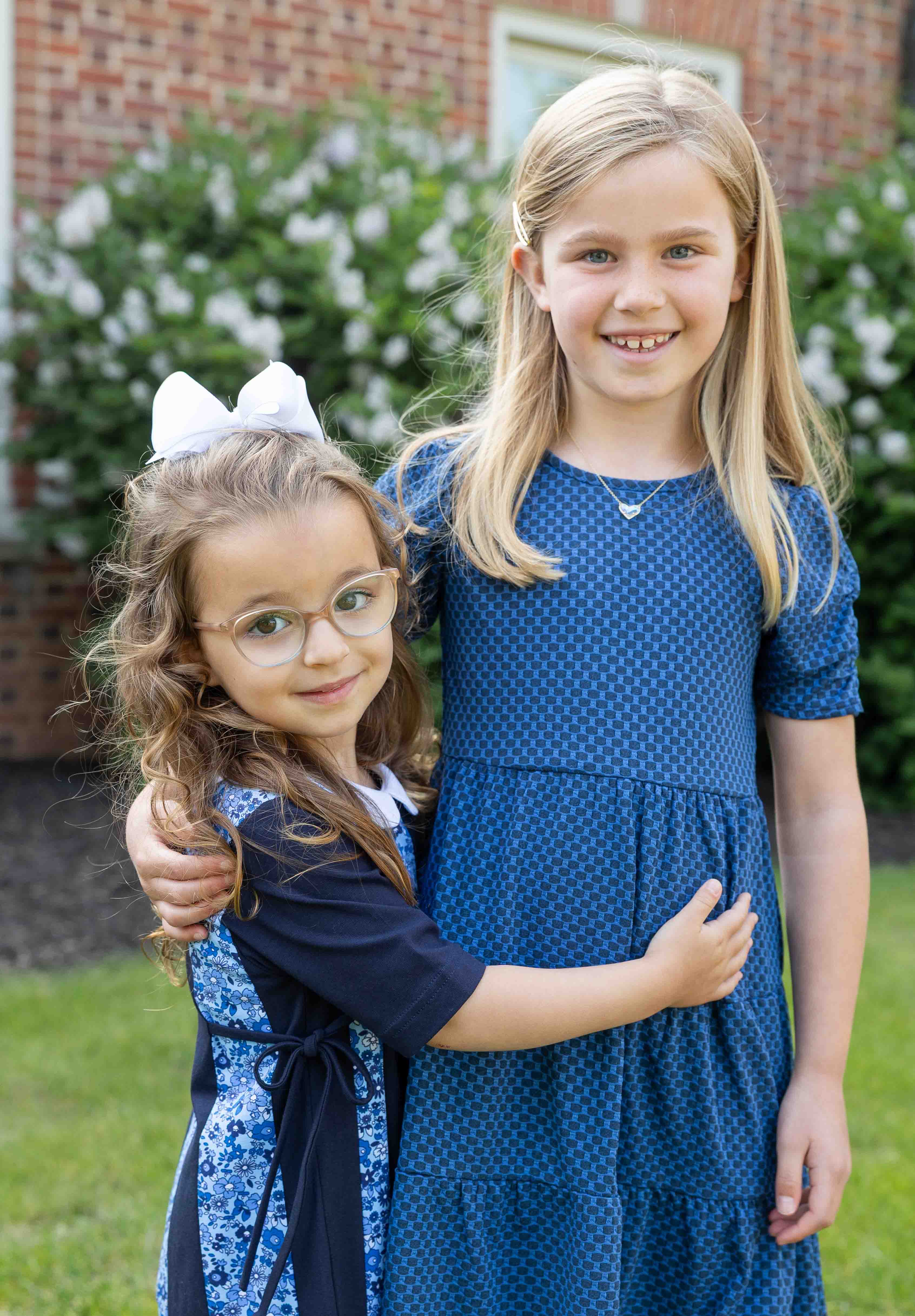 Two young girls in blue dresses standing outdoors with a brick building and greenery in the background.