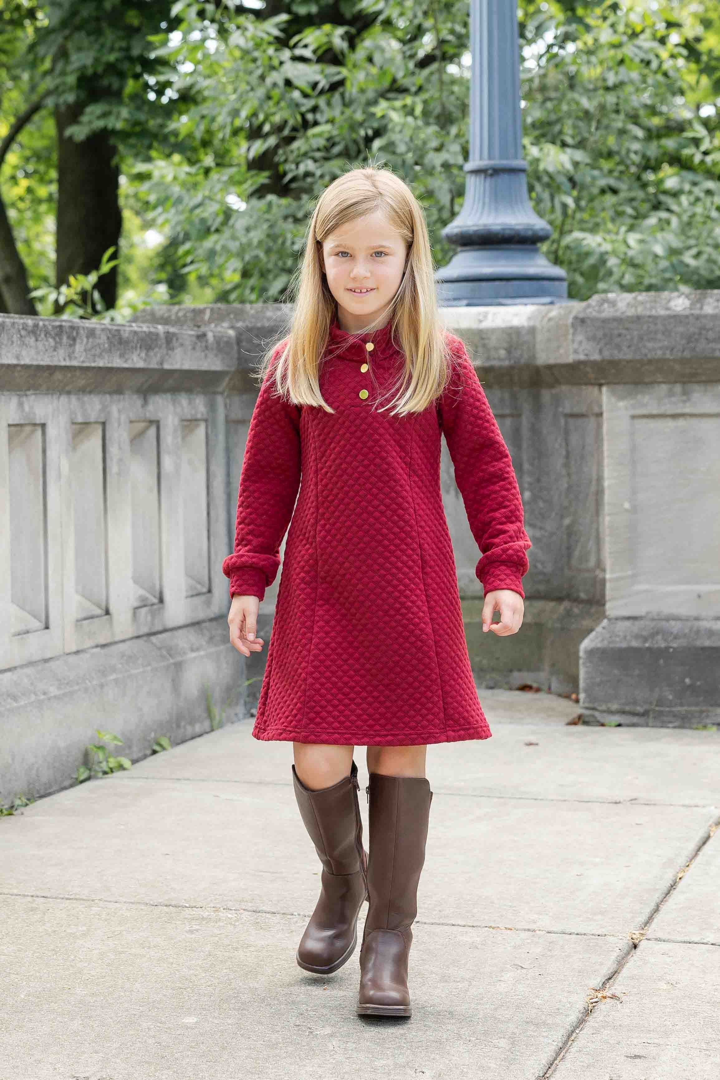 Young girl in a red dress and brown boots standing outdoors on a sidewalk.