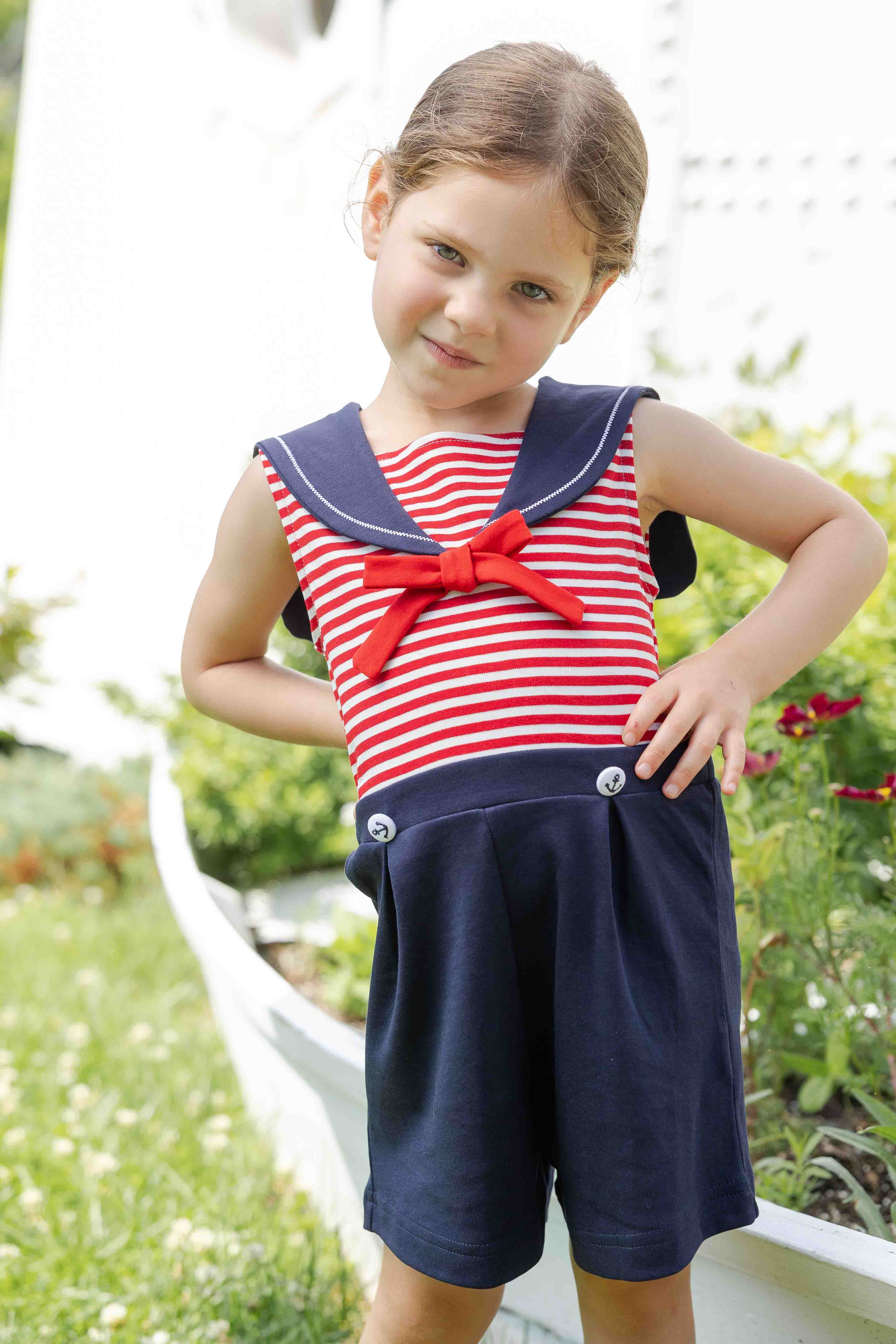 Young girl wearing a red and white striped dress with a navy blue bow and shorts, standing outdoors.