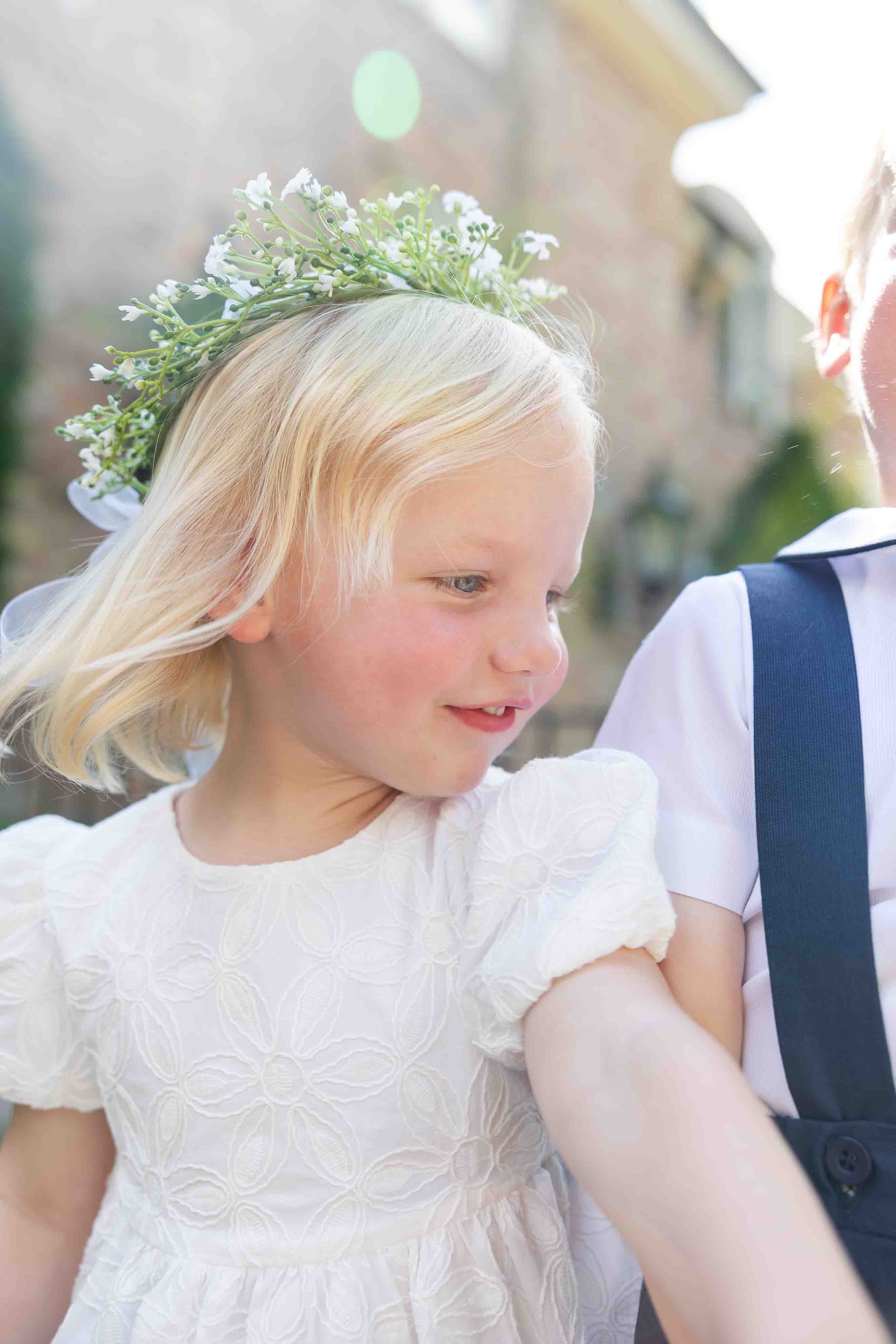 Young girl in a white dress with a floral headband, standing outdoors.