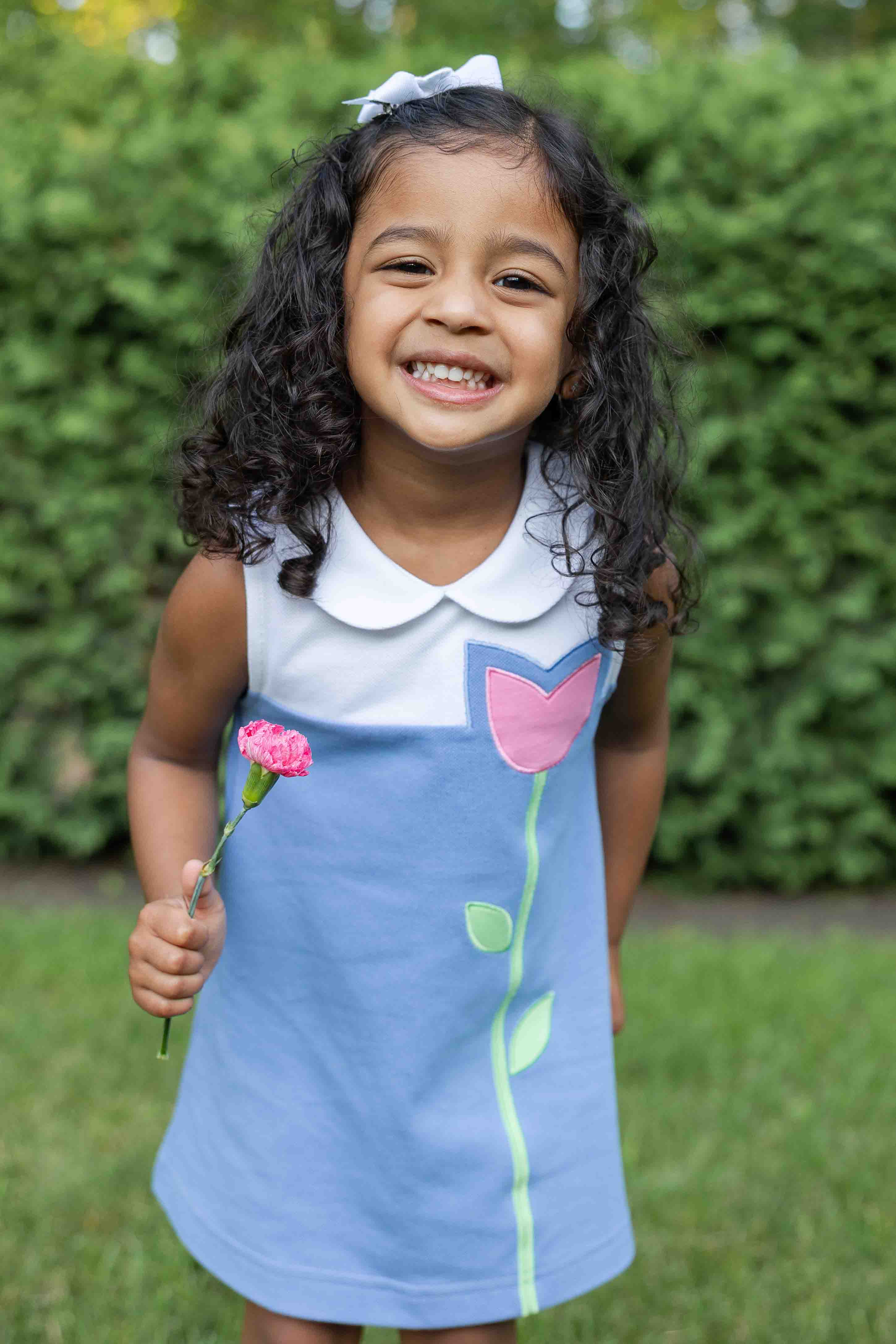 Young girl in a blue dress with floral design holding a pink flower, standing in front of green bushes.