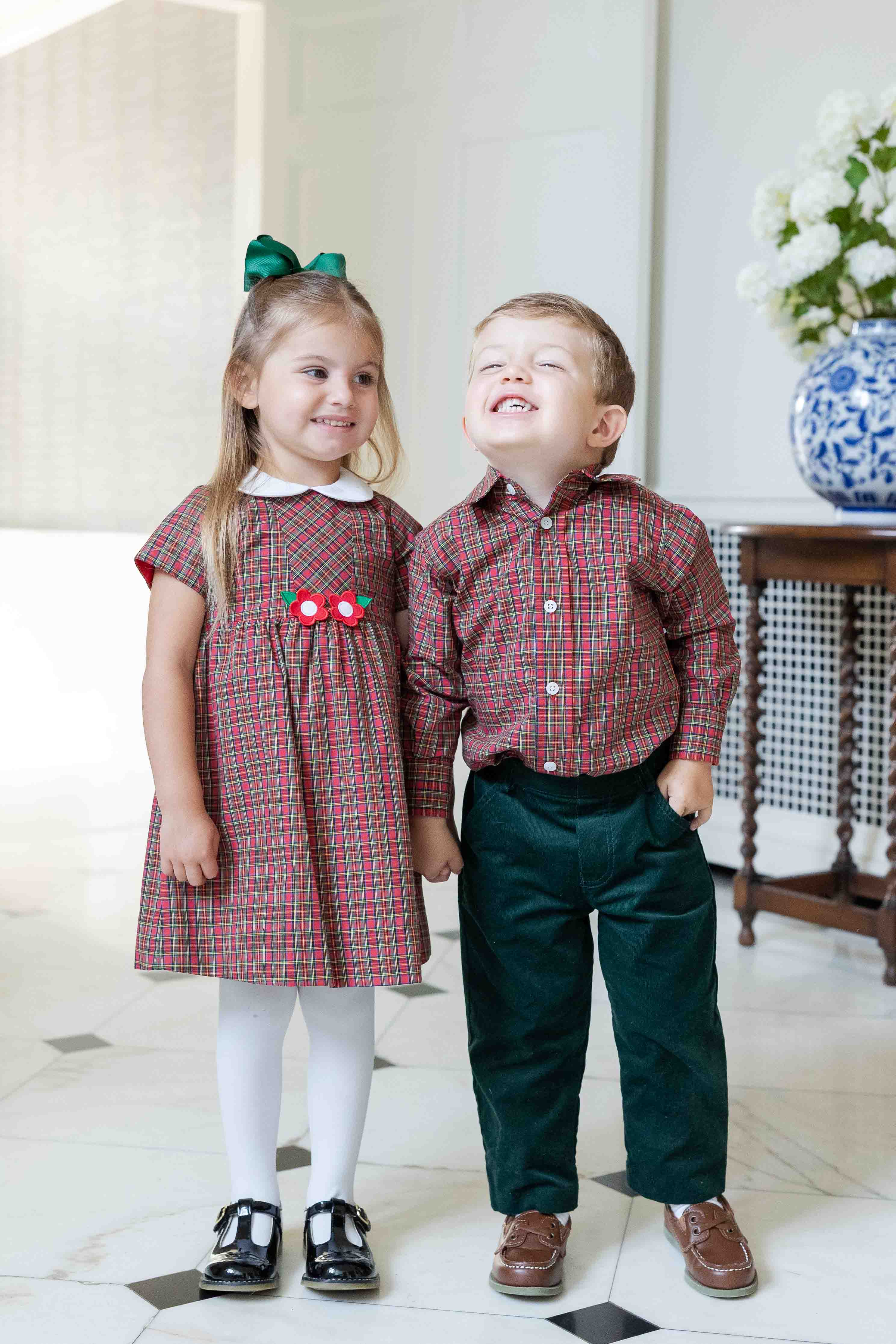 Two children in matching plaid outfits standing indoors.