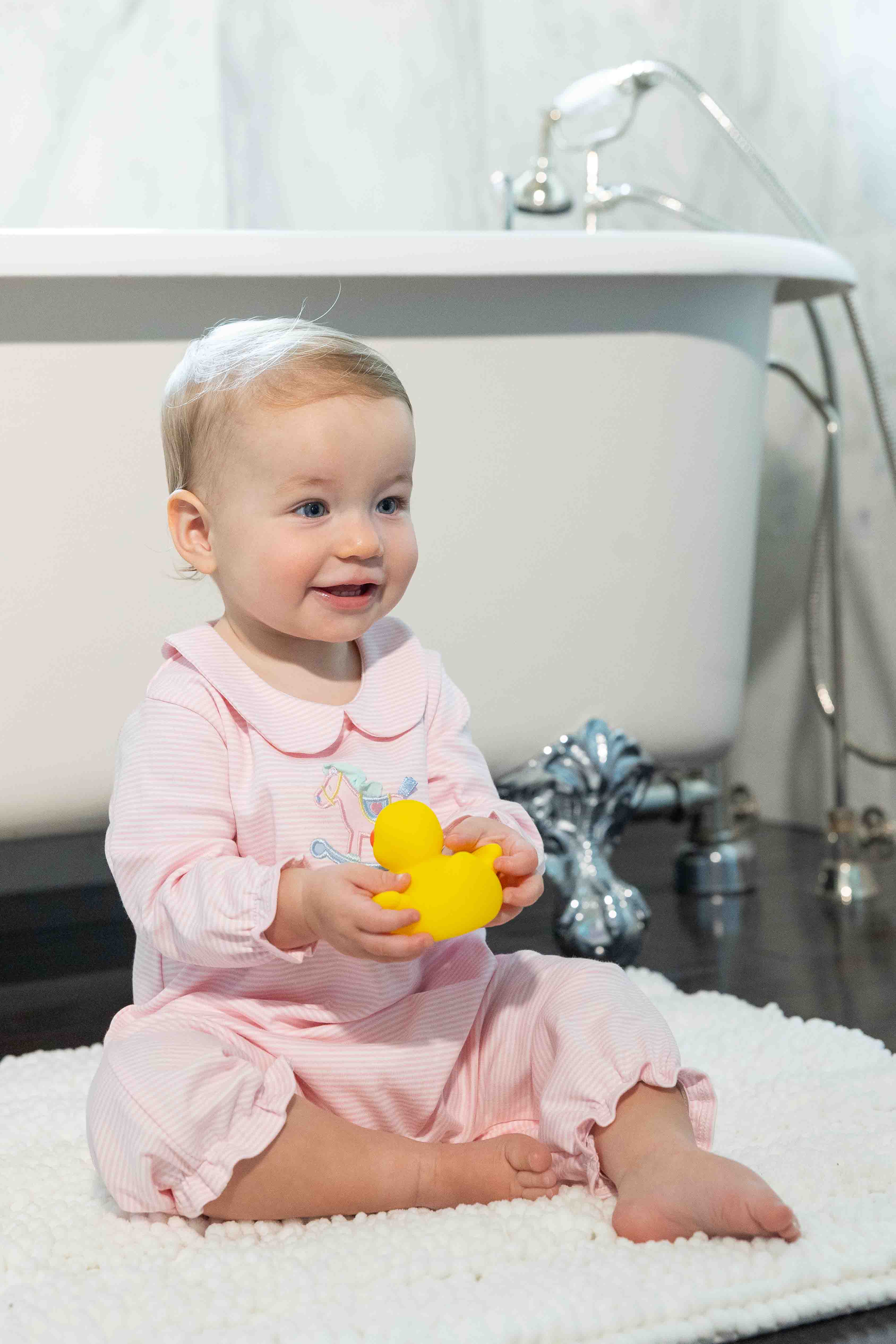 Baby in pink outfit holding a yellow rubber duck in front of a bathtub.