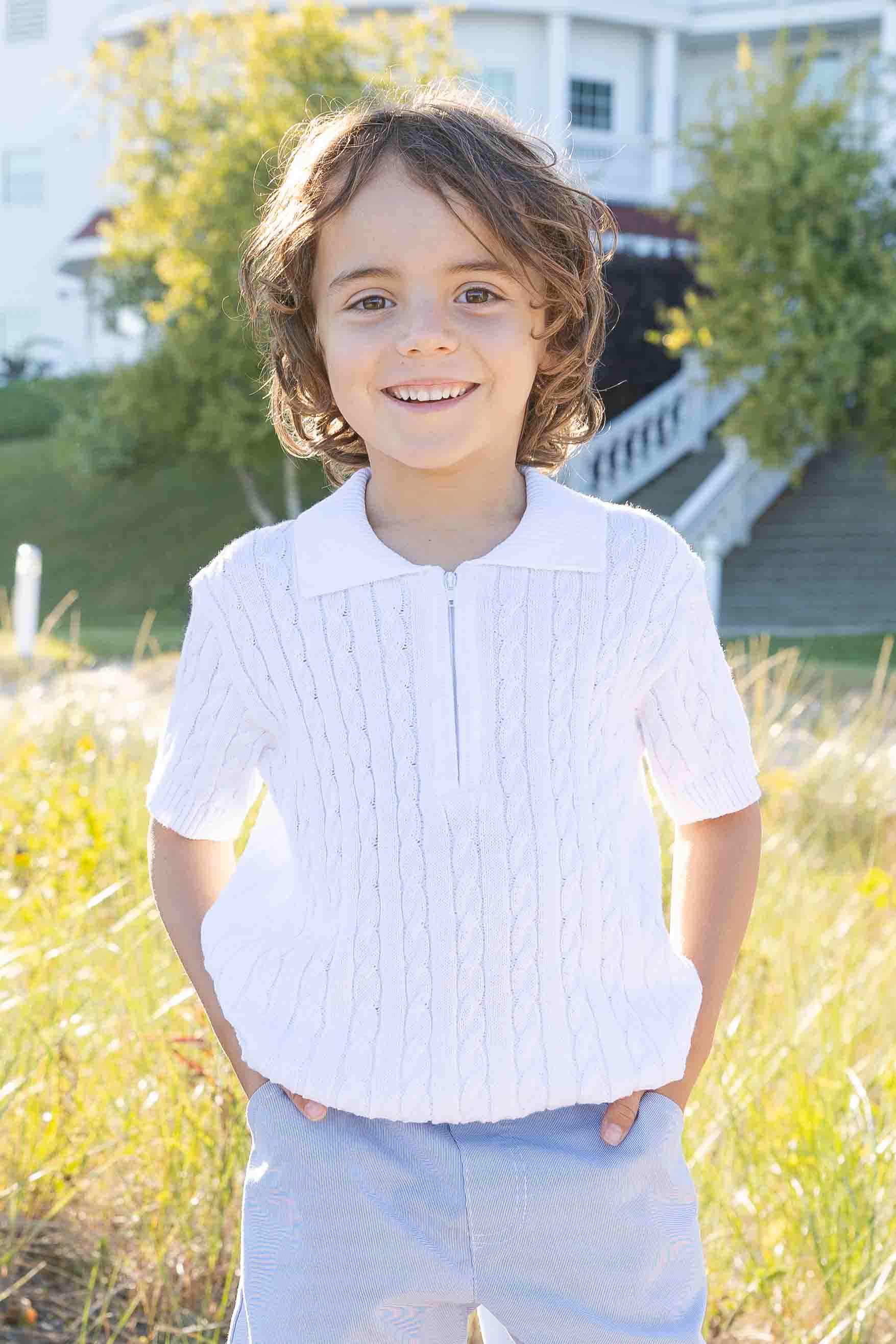 Child wearing a white shirt and light blue pants standing outdoors with greenery and a house in the background.