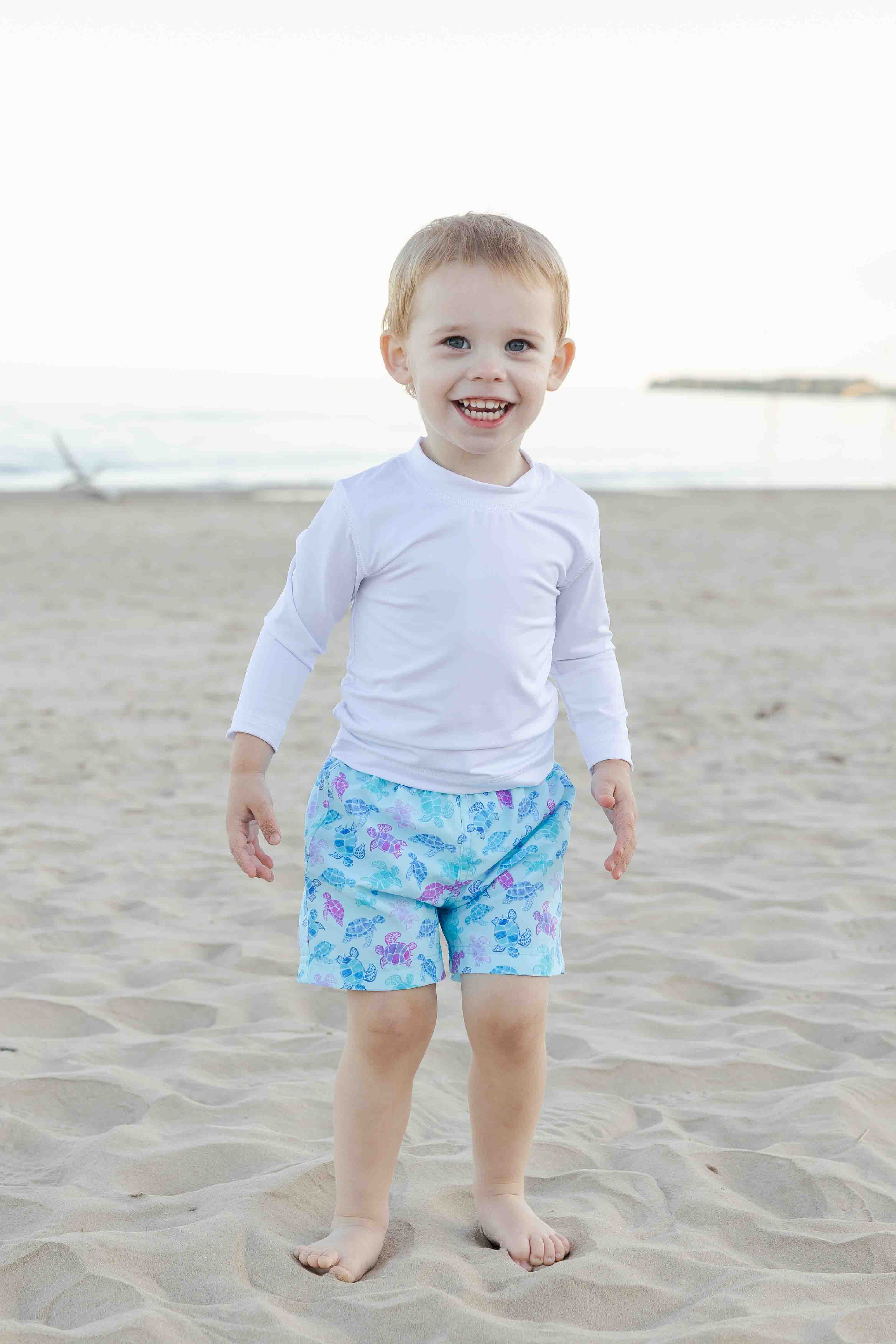 Child wearing a white long-sleeve shirt and floral shorts on a sandy beach.