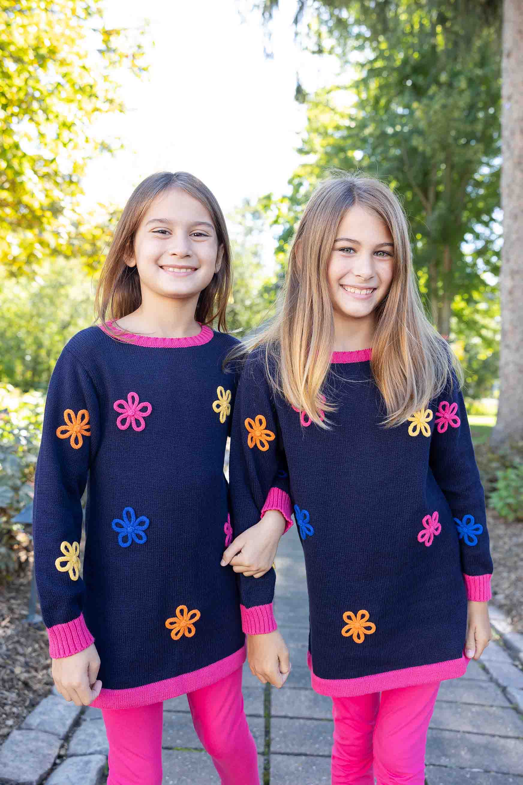 Two young girls wearing matching navy blue sweaters with colorful flower patterns outdoors.