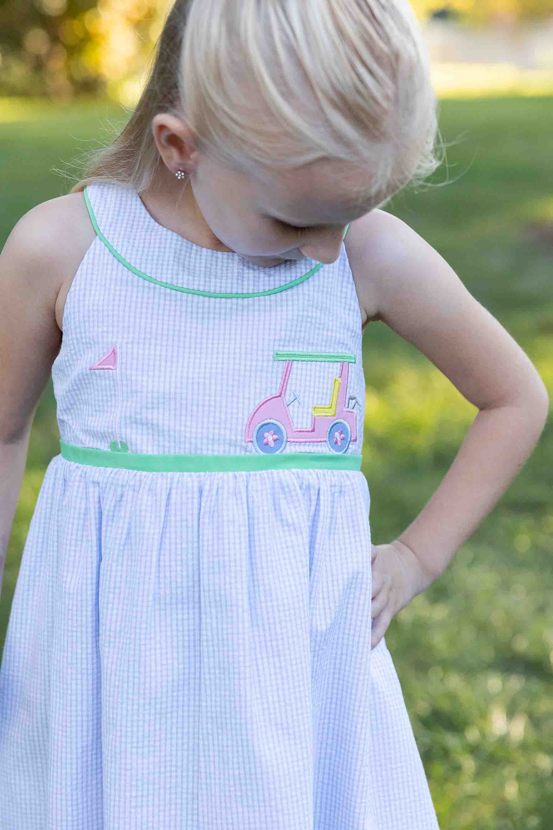 Young girl wearing a light blue dress with pink and green accents, standing outdoors.
