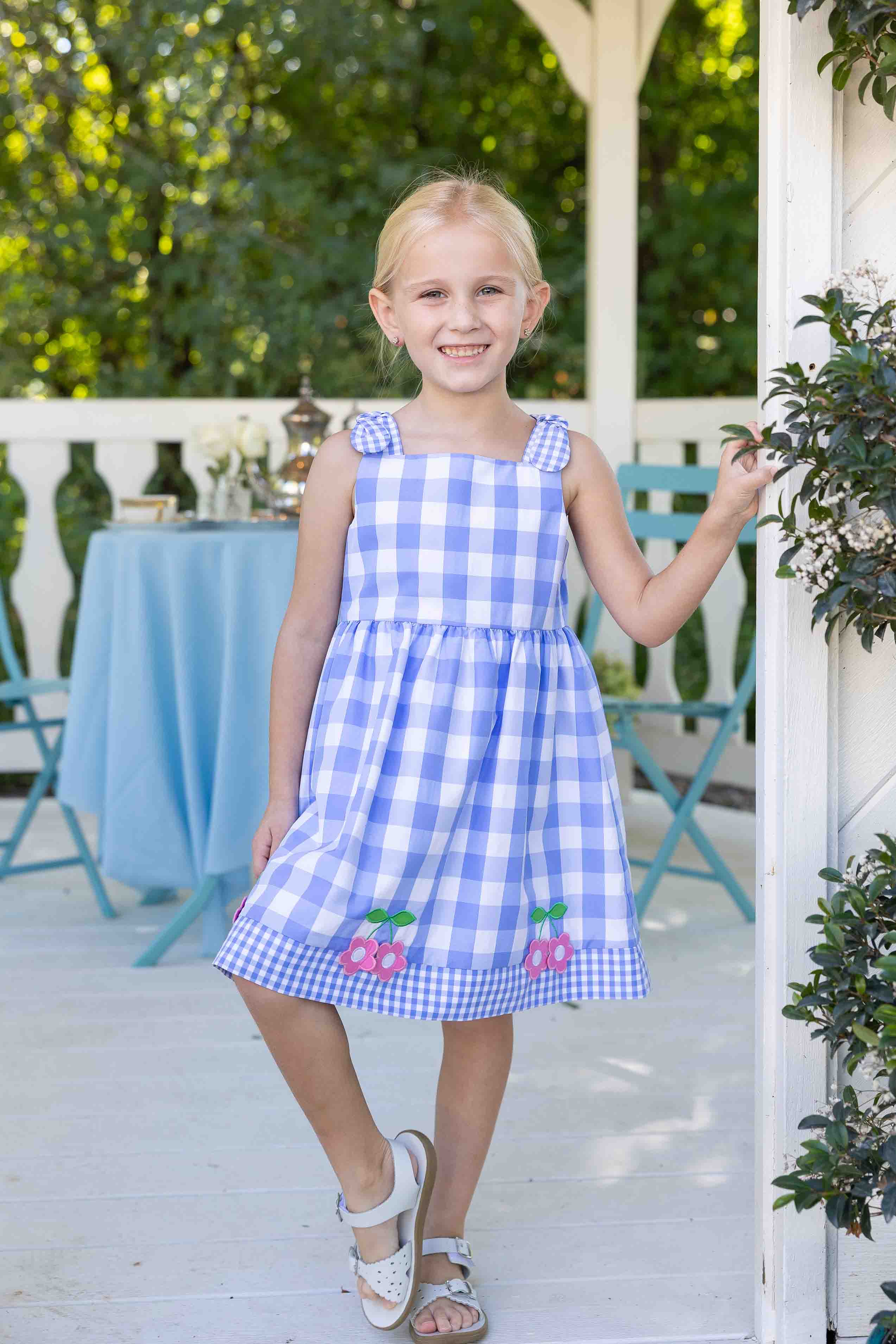 Young girl in a blue and white checkered dress standing on a porch.