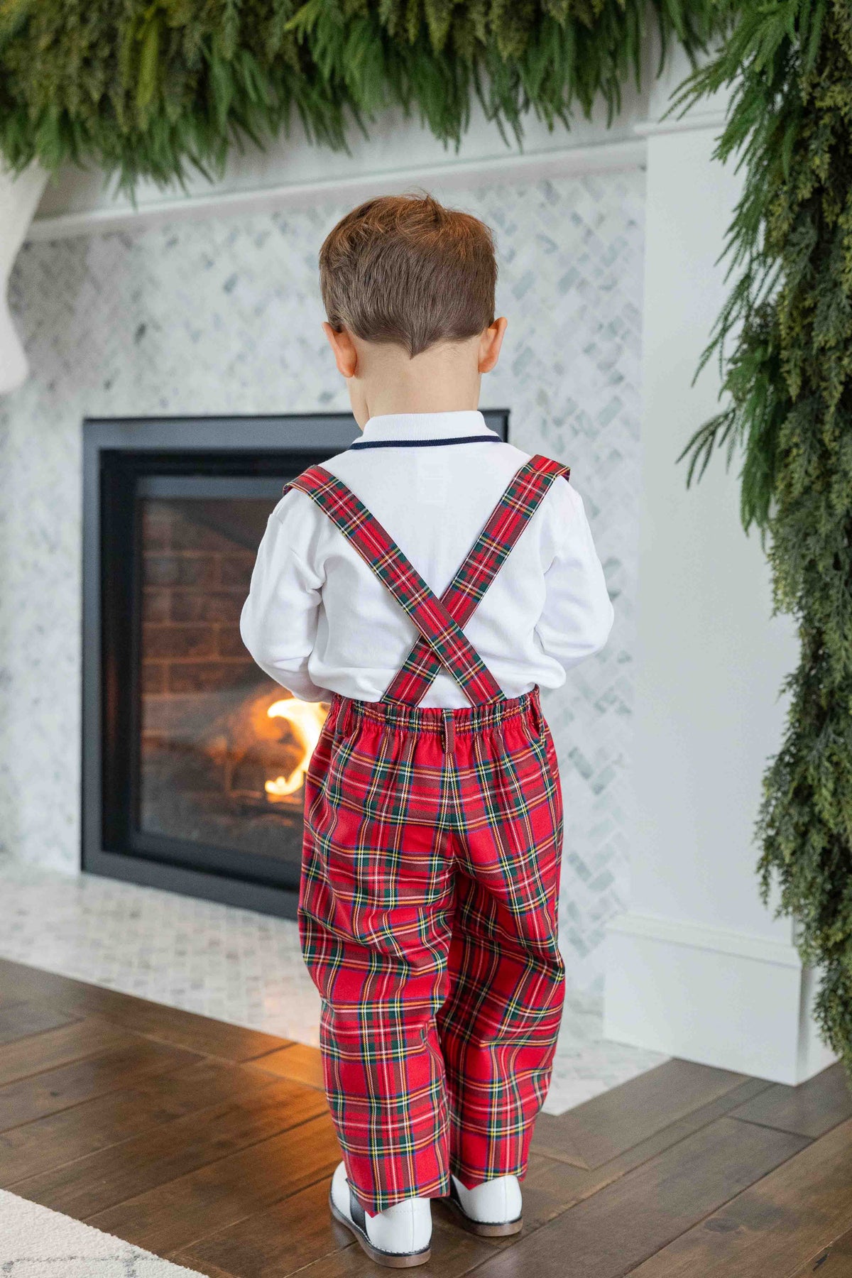 A toddler boy viewed from behind, wearing a white long-sleeve shirt and red tartan plaid suspenders with matching plaid trousers, standing in front of a fireplace.