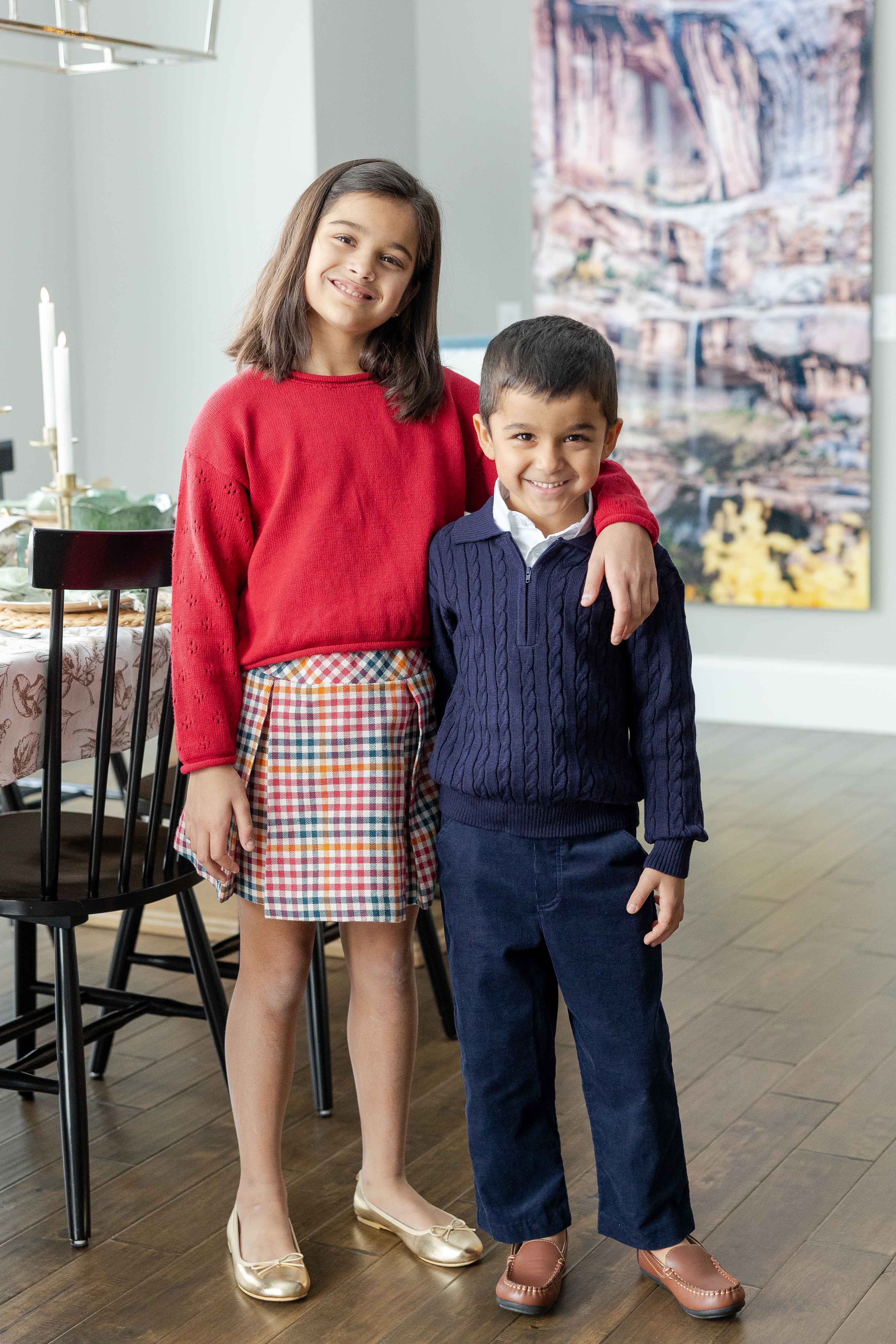 Two children standing together in a room with a painting on the wall.