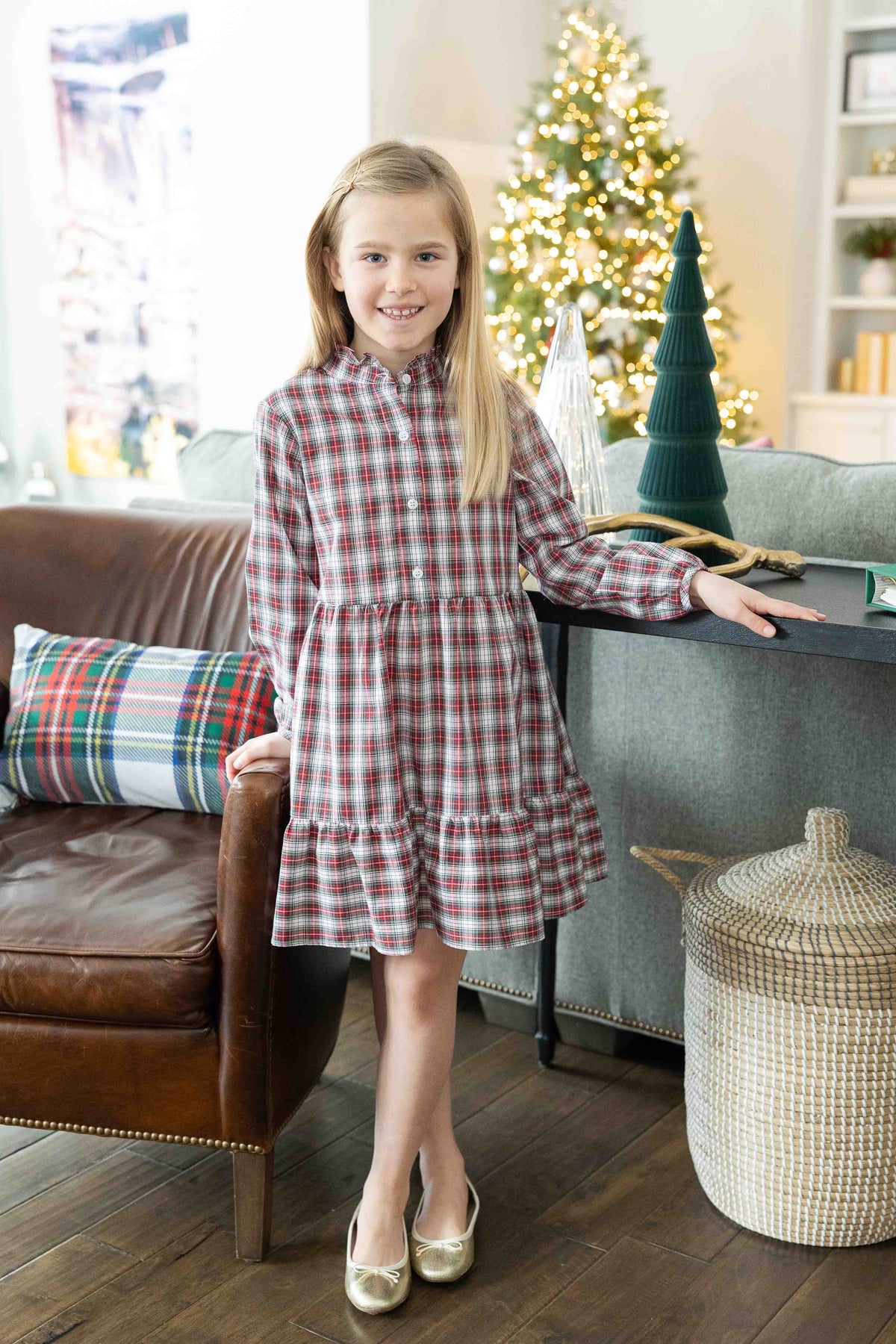 Young girl in a plaid dress sitting on a couch in a living room with a Christmas tree in the background.
