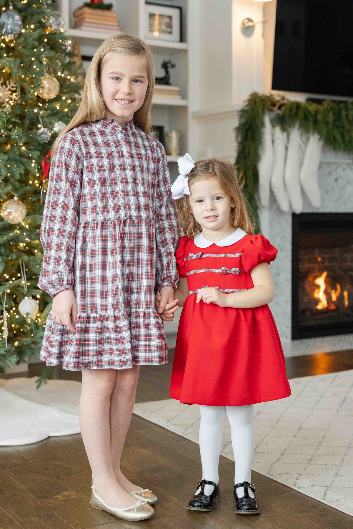 Two young girls in dresses standing in a festive living room with a Christmas tree and fireplace.