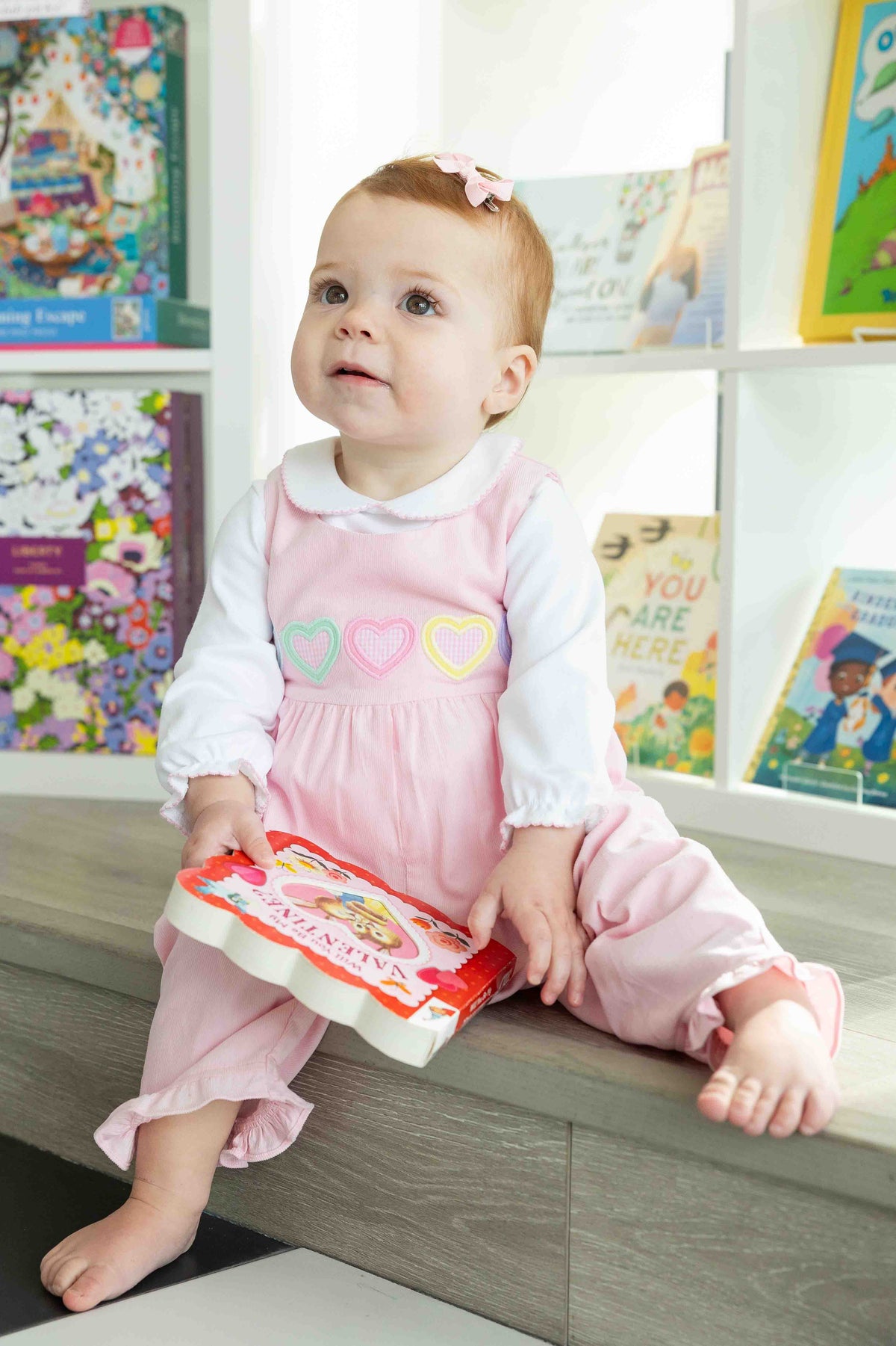 Baby in a pink dress sitting on a shelf with books in the background