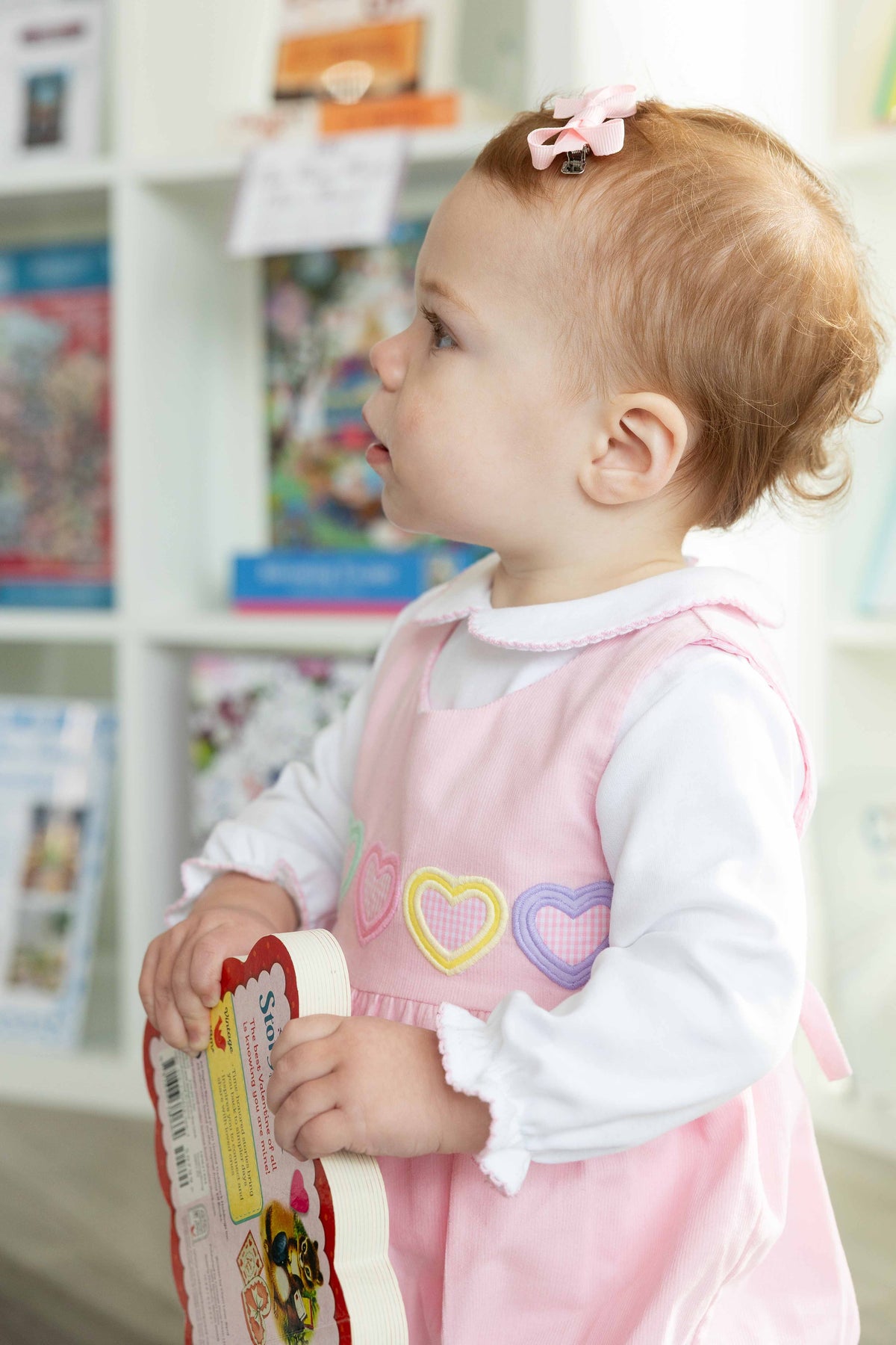 Child in a pink dress with heart patterns holding a book in a library setting.
