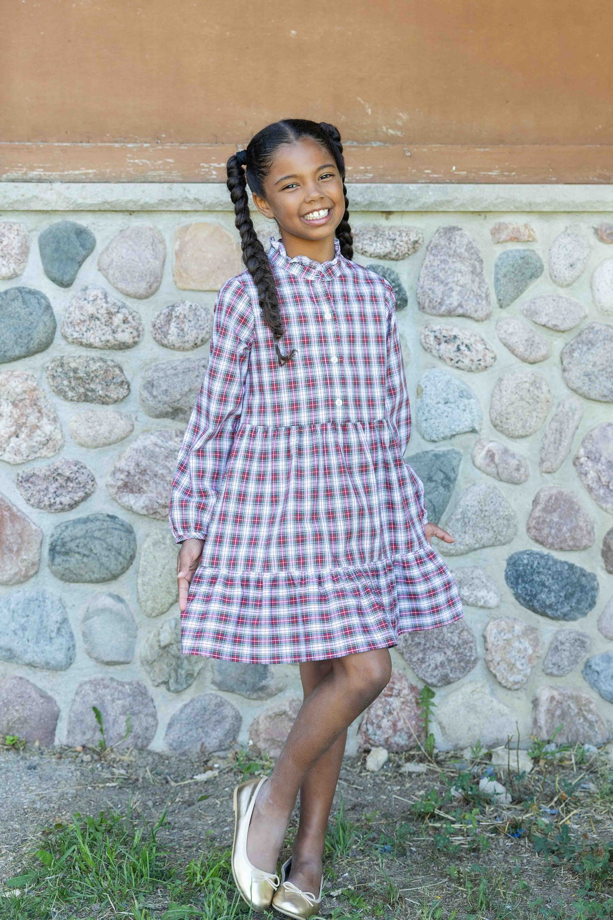 A young girl with braided hair wearing a red and white plaid long-sleeve dress, standing in front of a stone wall.