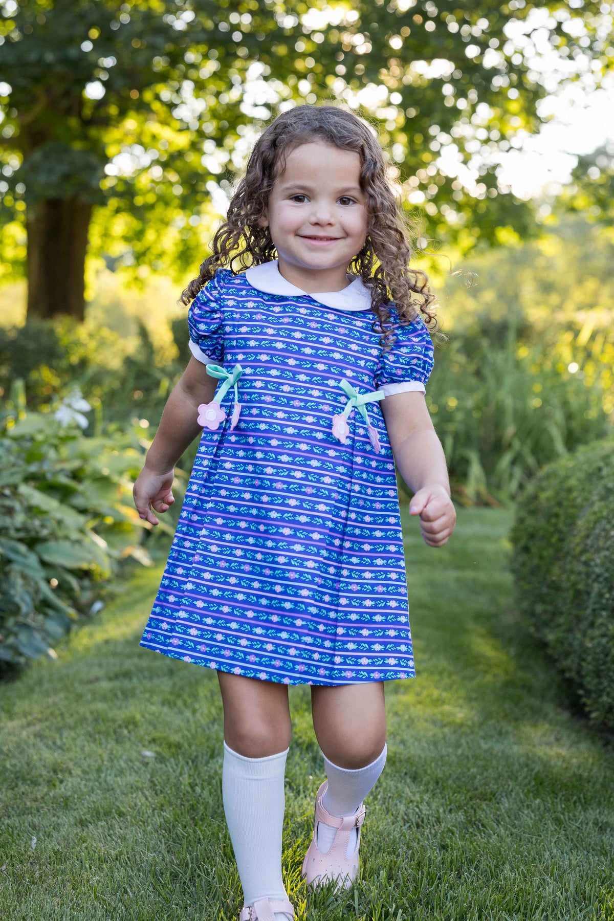 young girl in a blue patterned dress with white collar walking in a garden