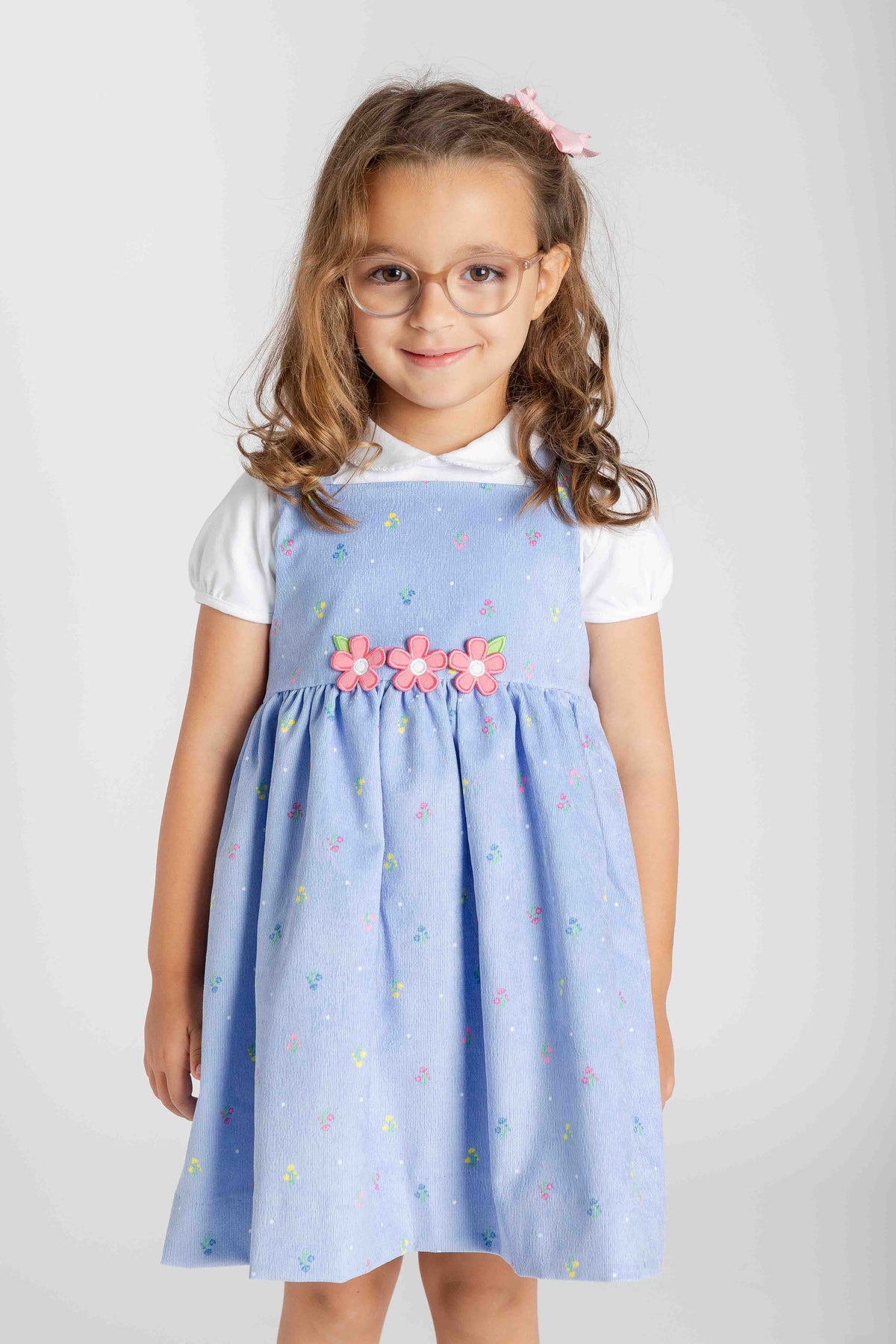 A young girl with glasses and a pink hair clip is wearing a light blue pinafore dress with a white short-sleeve shirt underneath. The dress features a smocked bodice, a waistband decorated with pink fabric flowers, and a flared skirt with a subtle pastel pattern.