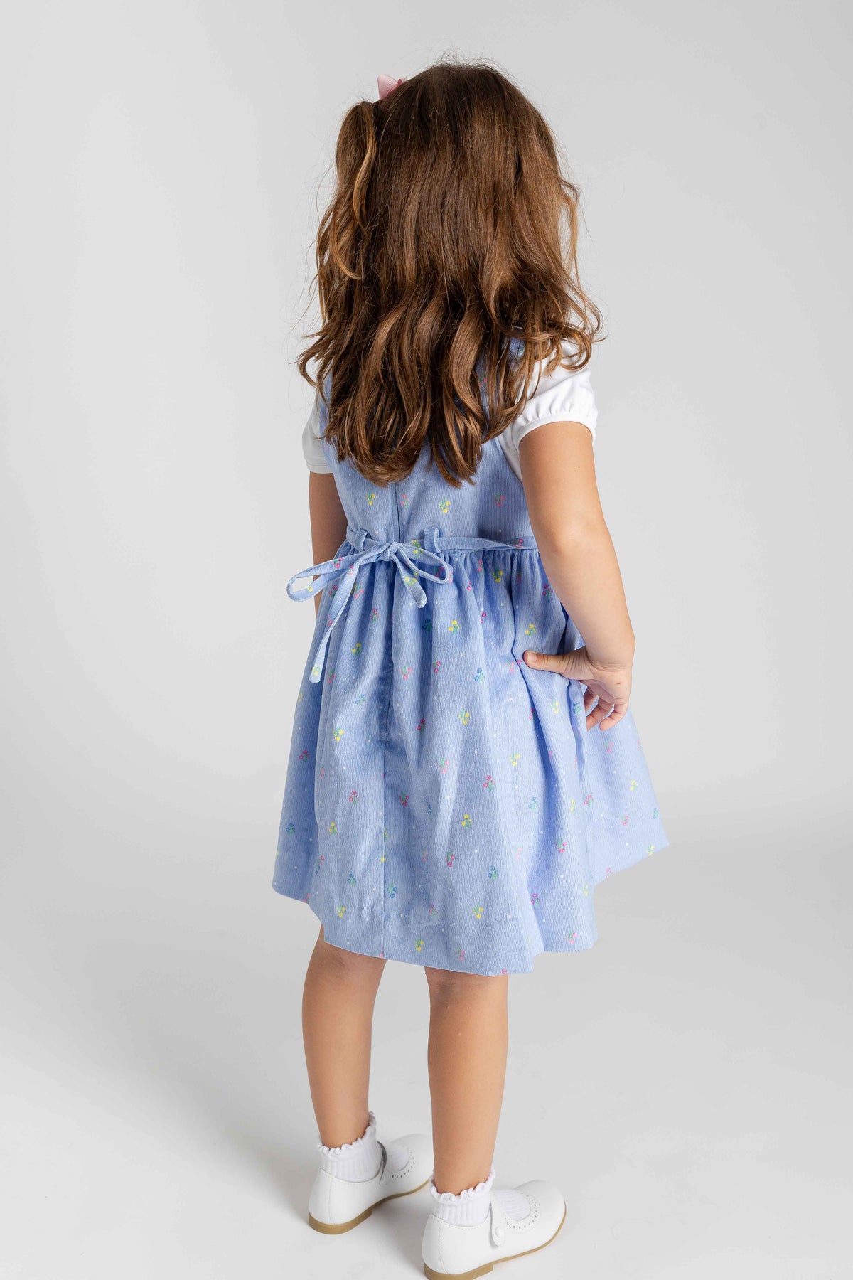 A young girl with long brown hair is shown from behind wearing a light blue pinafore dress with a tiny multicolor pattern, a white t-shirt, white socks, and white shoes, photographed against a neutral light gray background.