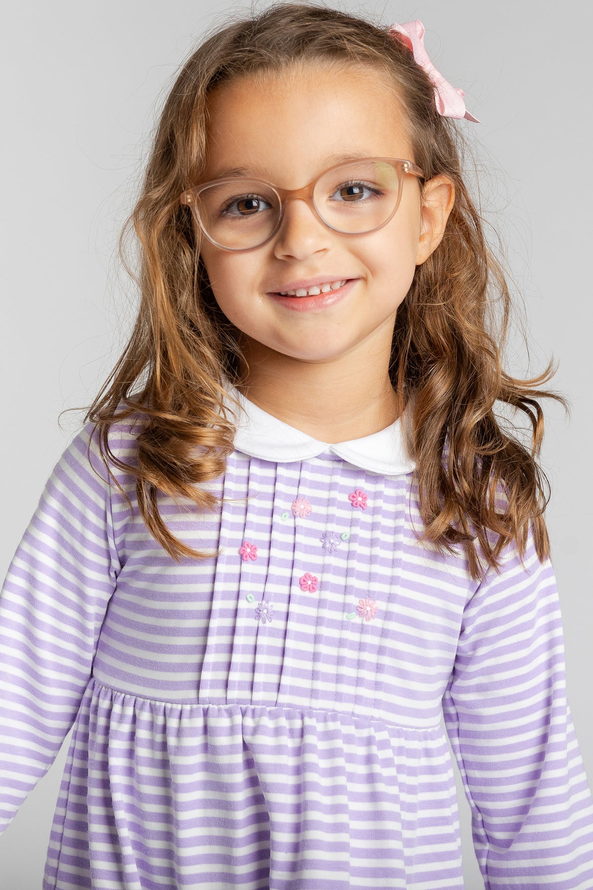 A young girl with long wavy brown hair and glasses, wearing a lavender and white striped long-sleeve dress with a white Peter Pan collar and small pink embroidered flowers, photographed against a solid light gray background.