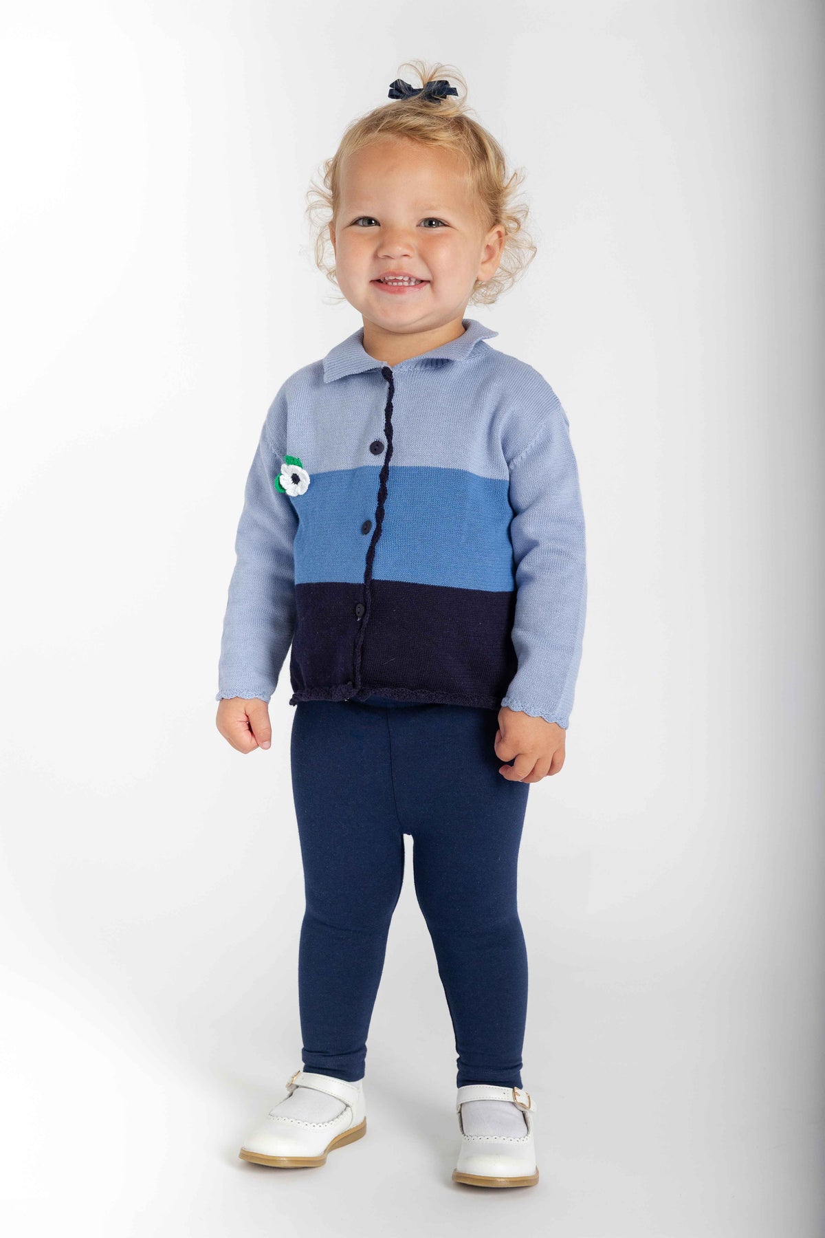A toddler girl stands against a light gray studio background wearing a color-block knit cardigan in light blue, medium blue, and navy, paired with navy leggings and white shoes.
