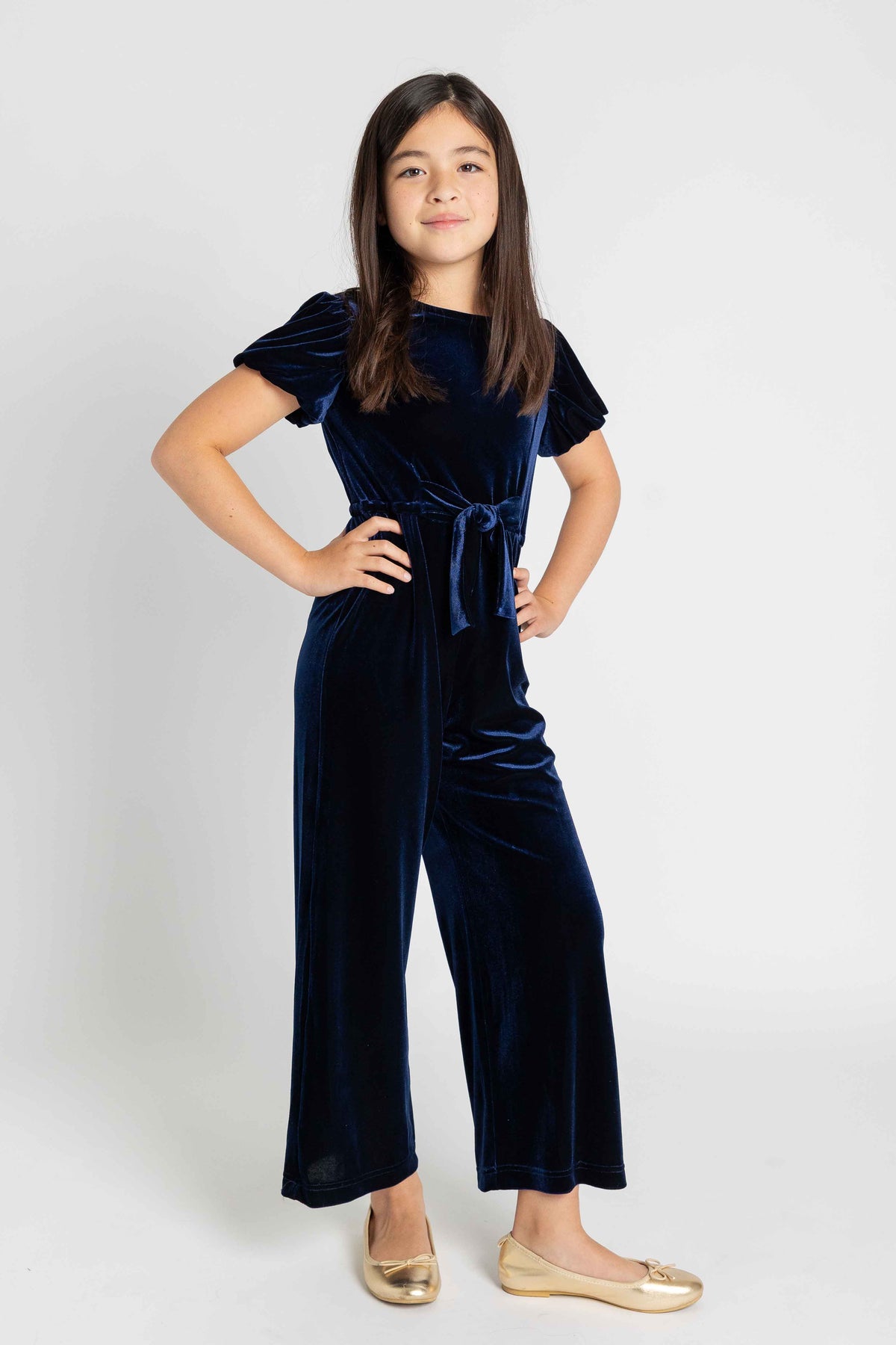 A young girl modeling a navy blue velvet short-sleeve jumpsuit with a tie waist, paired with gold flats, against a light gray studio background.