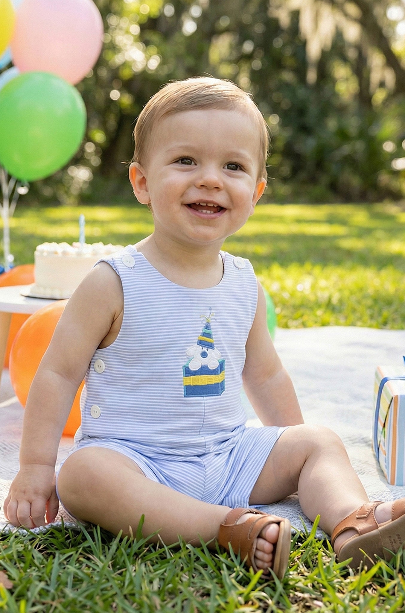 Child in a striped romper sitting outdoors with balloons and a cake in the background