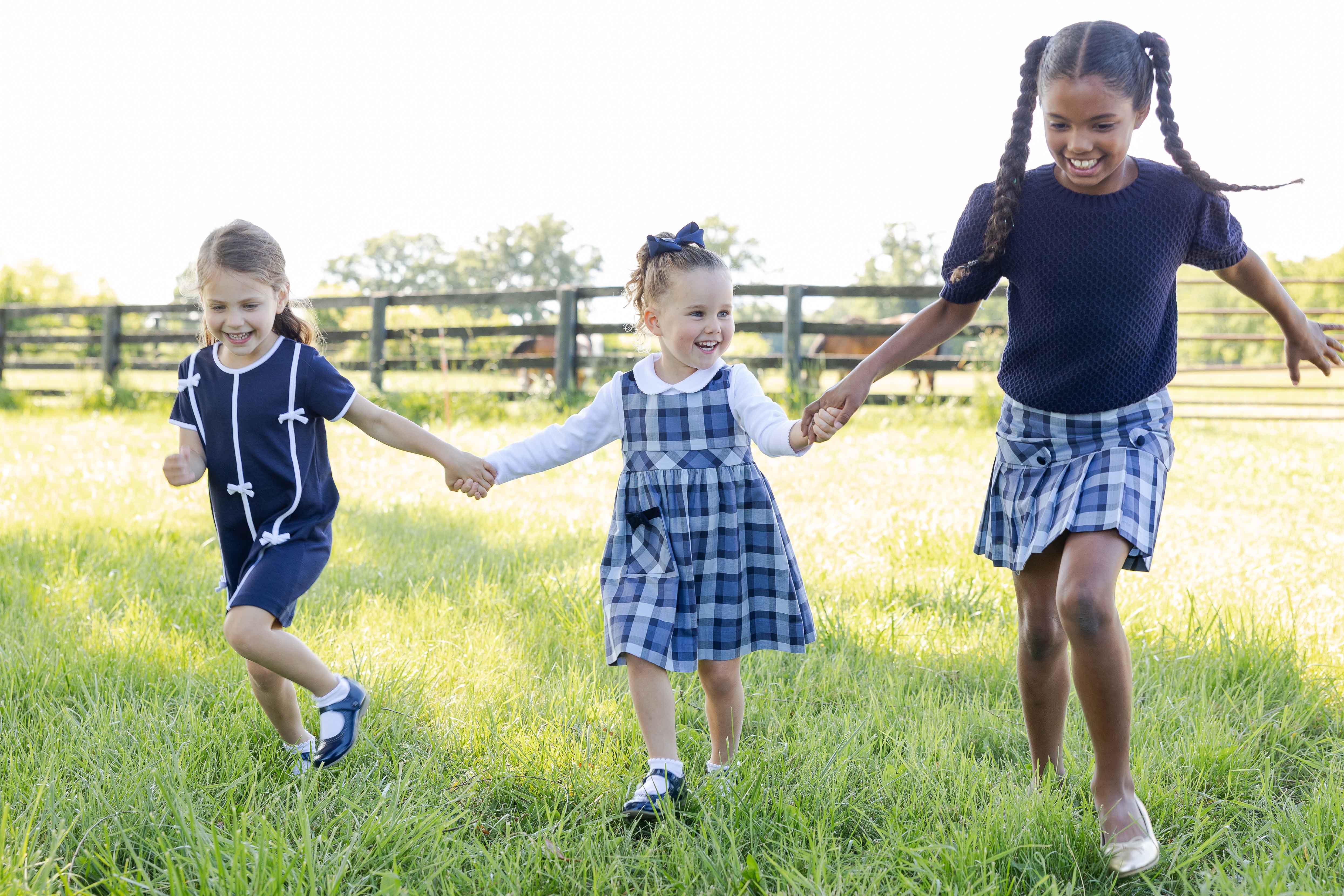 Three young girls in matching navy and blue plaid outfits run hand-in-hand across a sunny green grassy field.