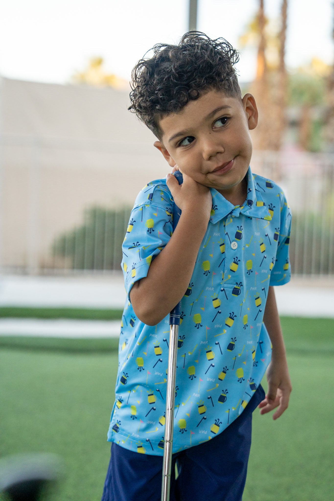Child in a blue shirt with a pattern, standing outdoors on a grassy area.