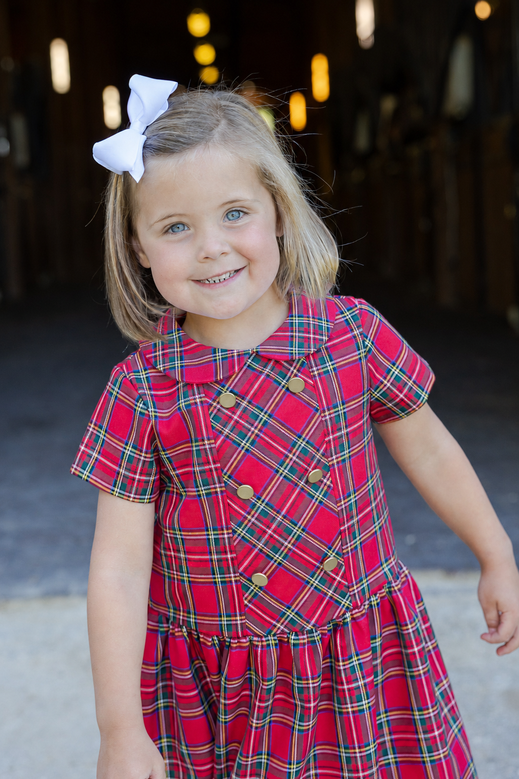 A young girl with a white bow in her hair is wearing a red plaid short-sleeve dress with gold buttons, standing in front of a blurred barn interior.