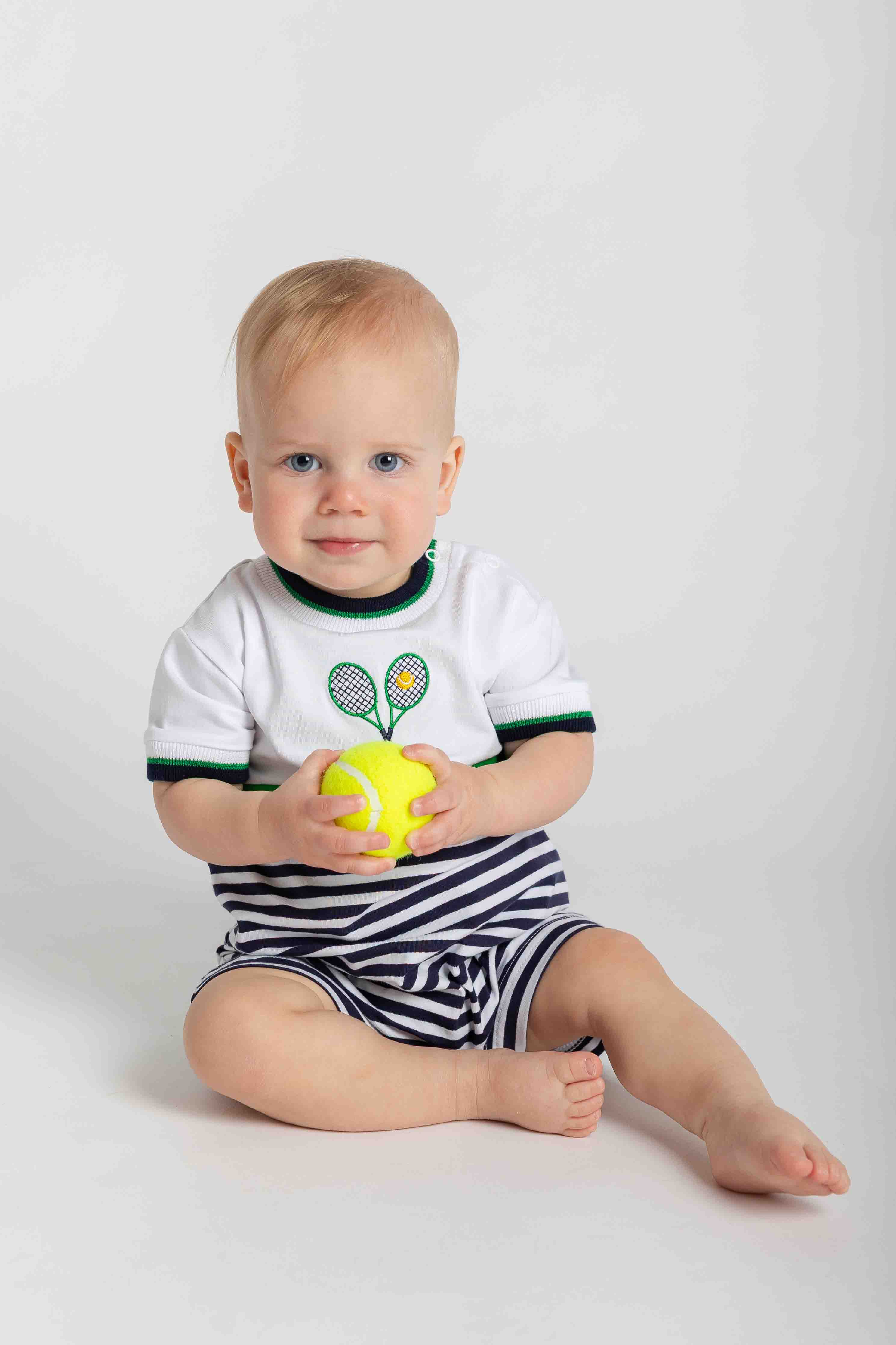 Baby sitting on a white background wearing a shortall, holding a yellow ball.