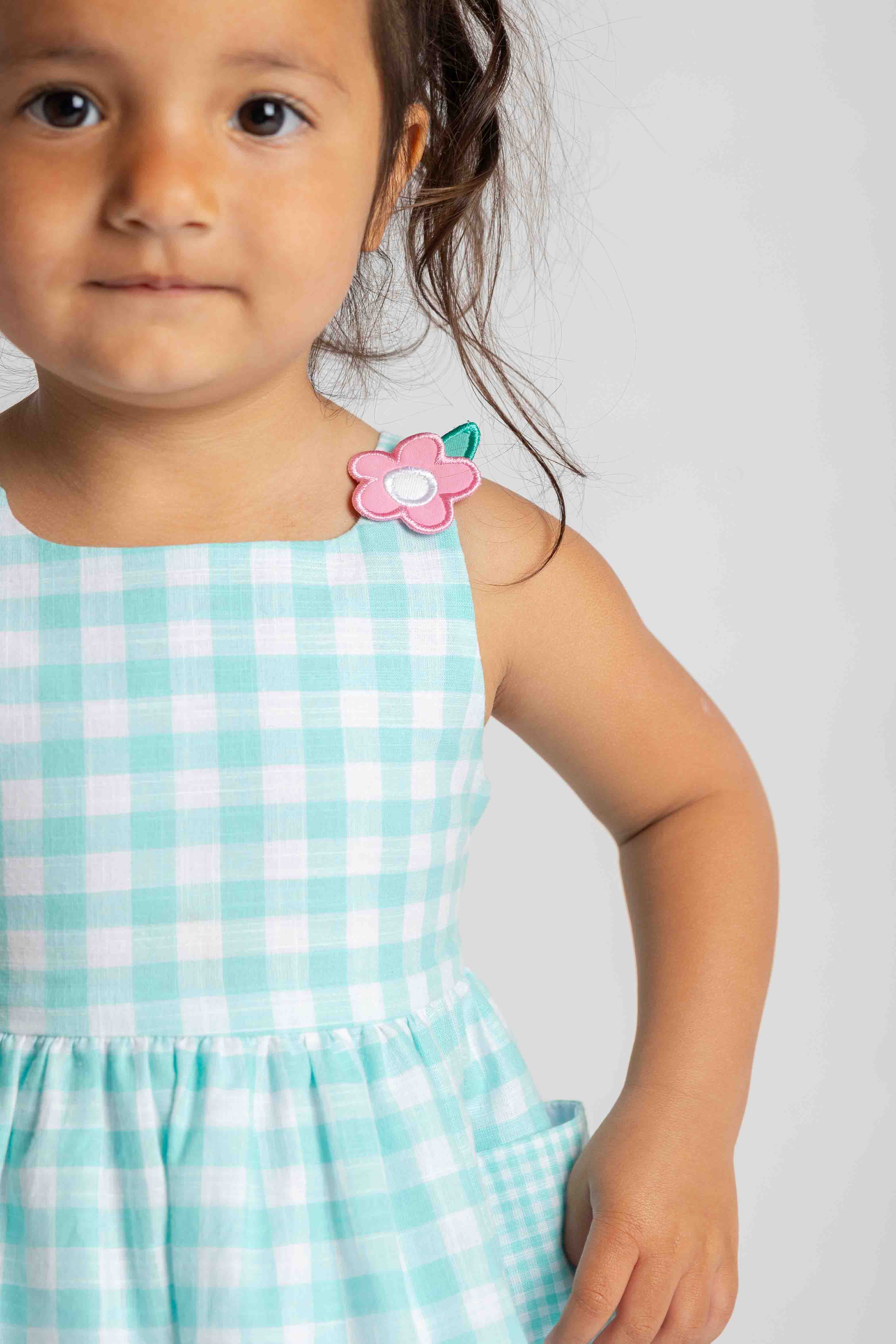 Child wearing a turquoise checkered dress with a pink flower accessory on a plain background