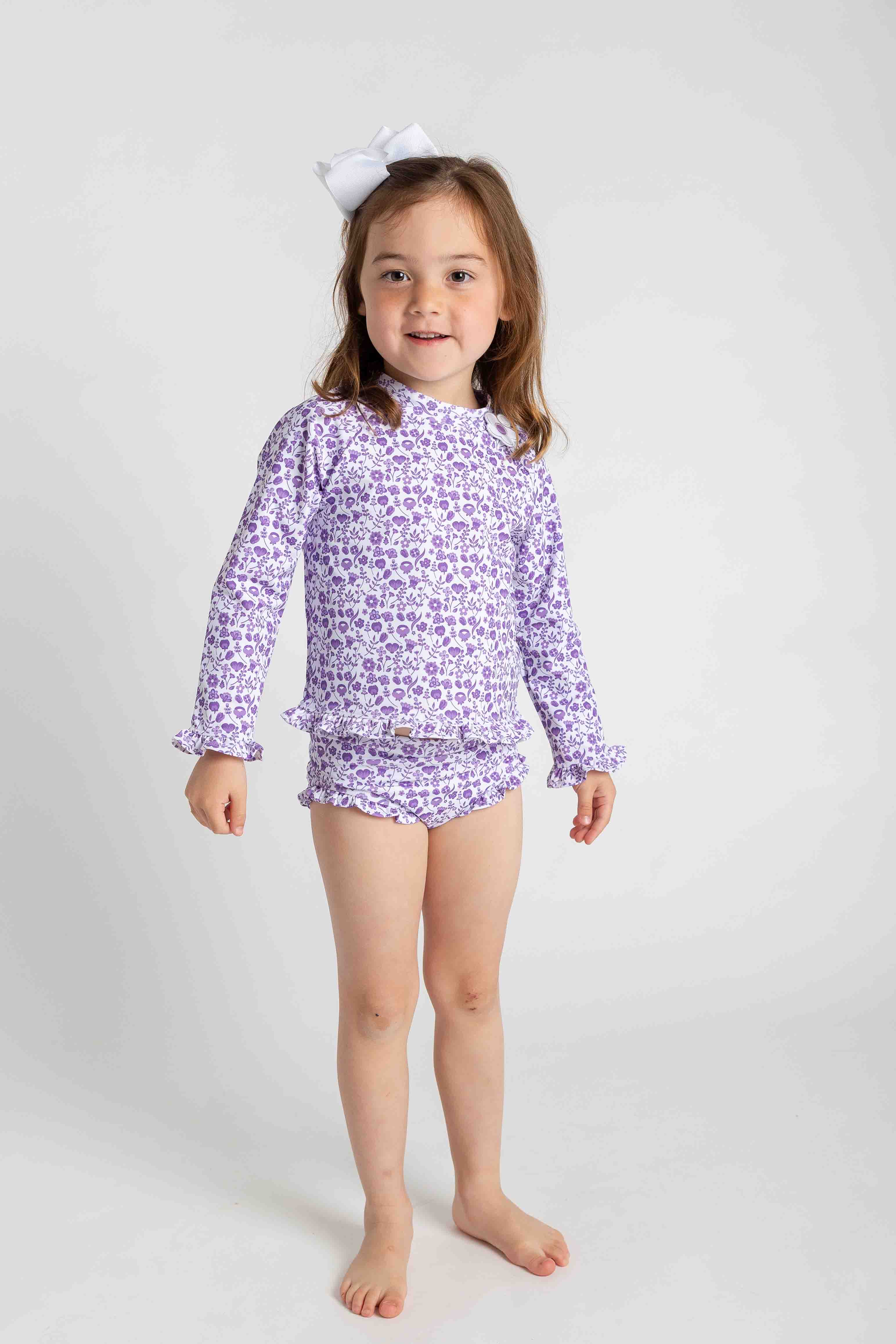 Young girl wearing a purple floral long-sleeve rash guard and matching shorts on a white background