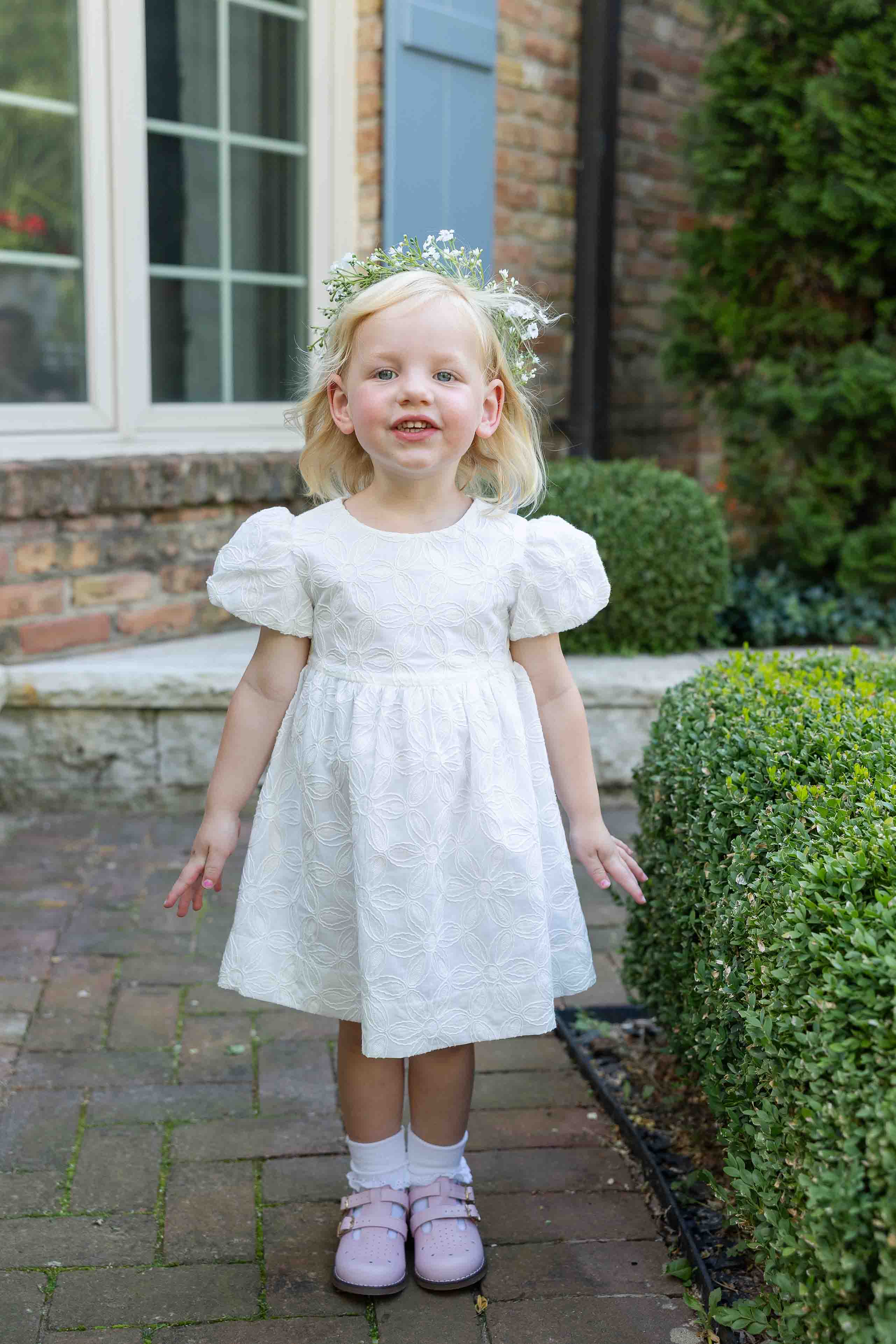 Young girl in a white dress standing outdoors with greenery and a building in the background