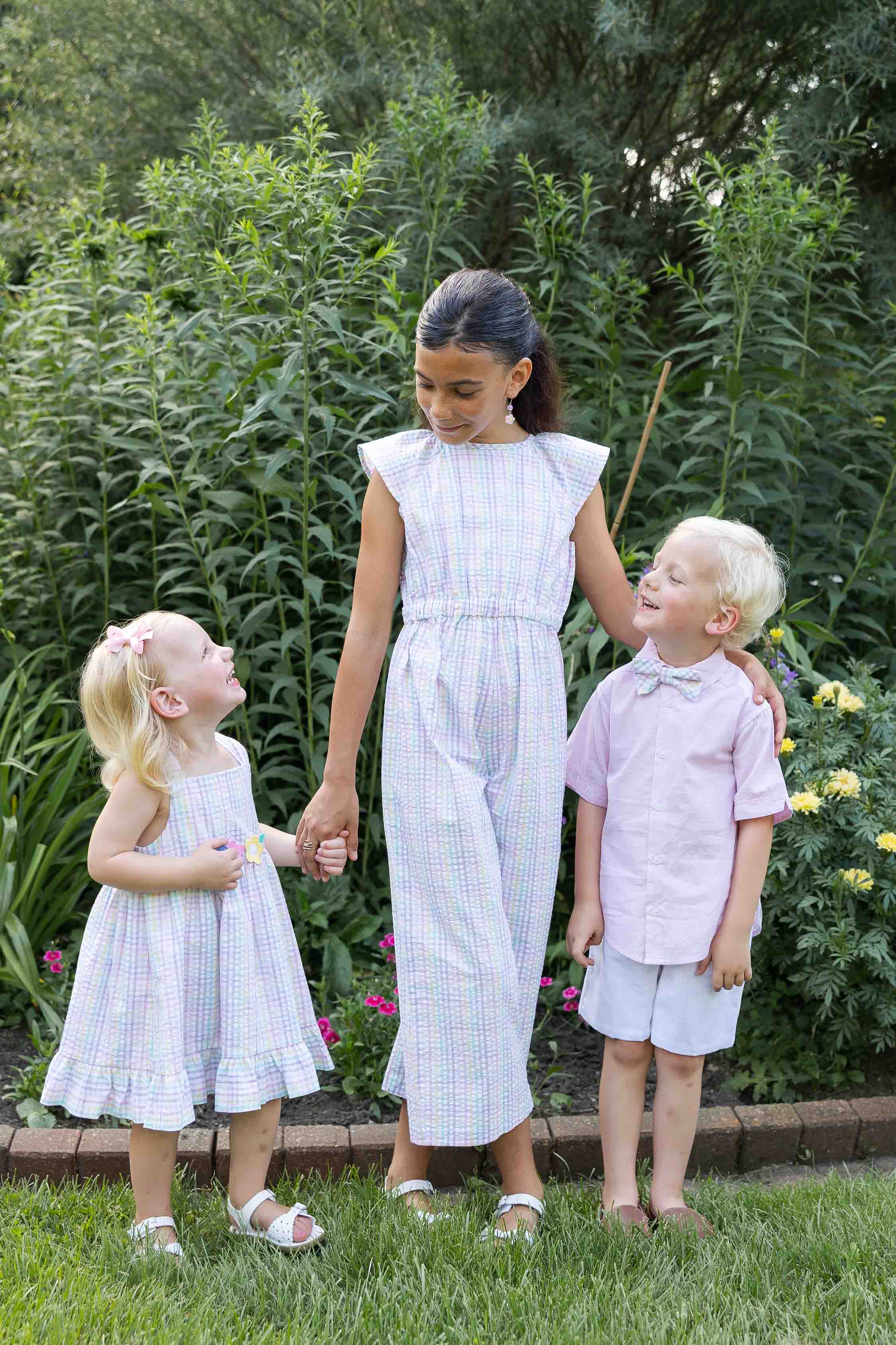 Three children in matching outfits standing outdoors with greenery in the background