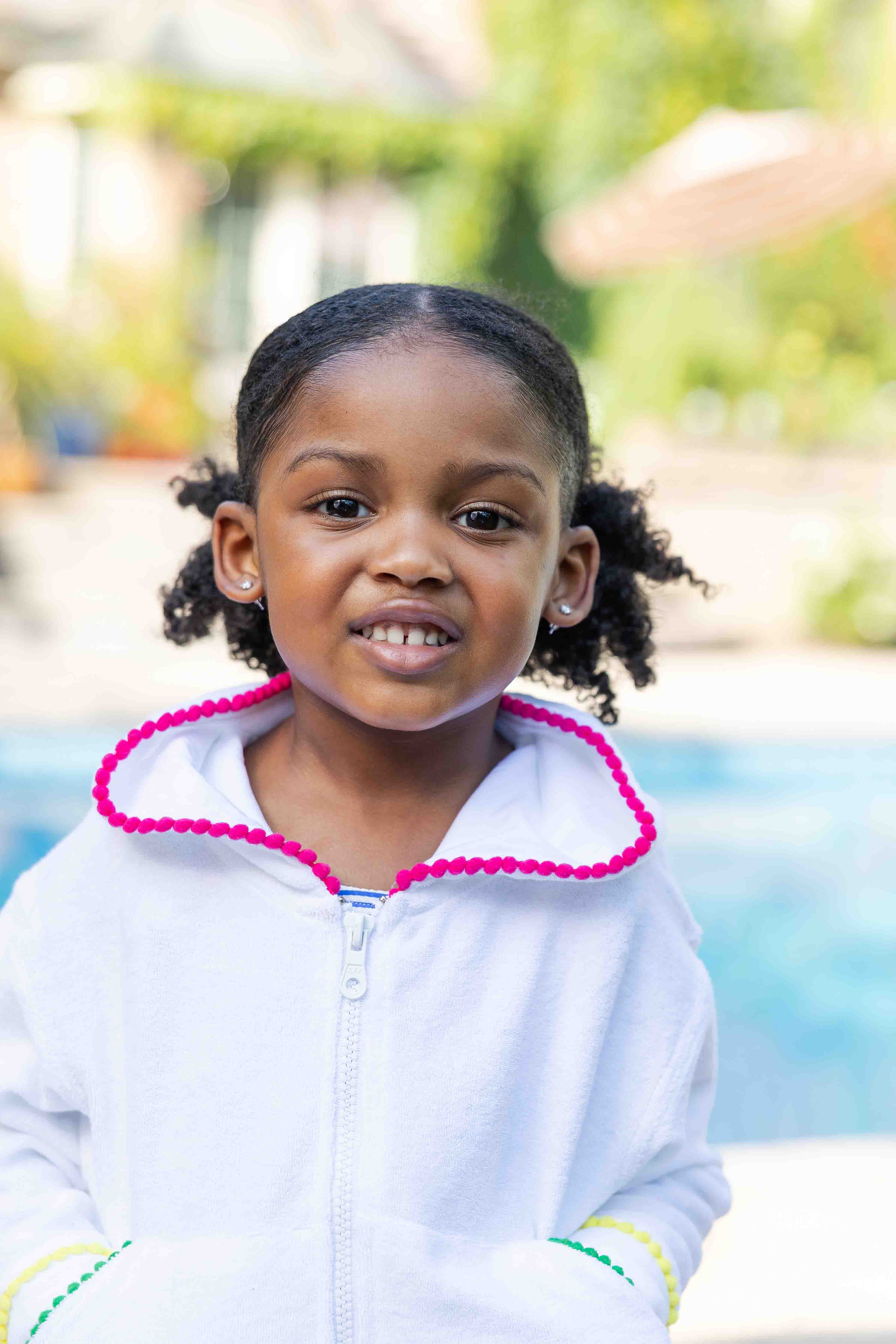 Young girl with braided hair wearing a white coverup with colorful trim, standing by a pool.