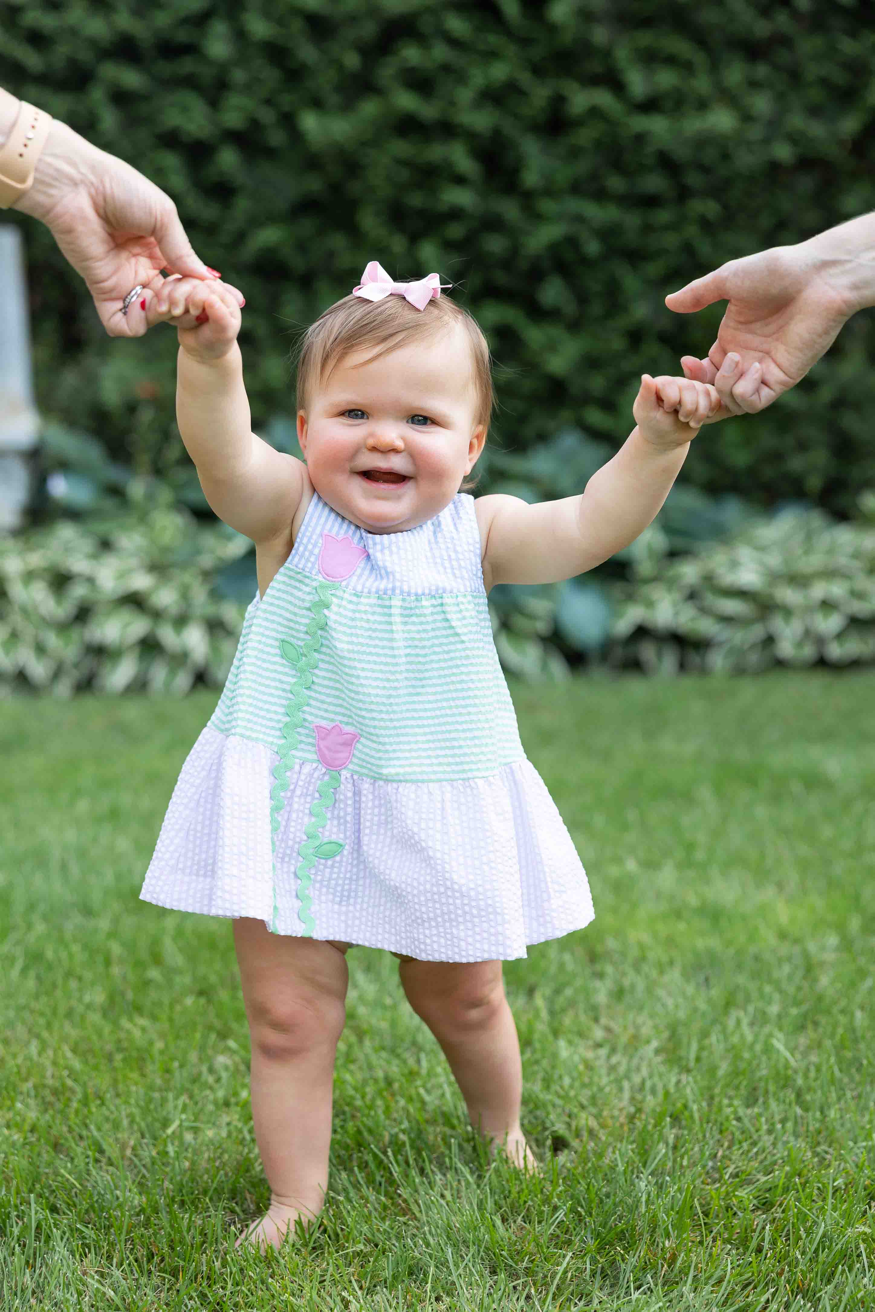Baby in a dress standing on grass with hands held by two people