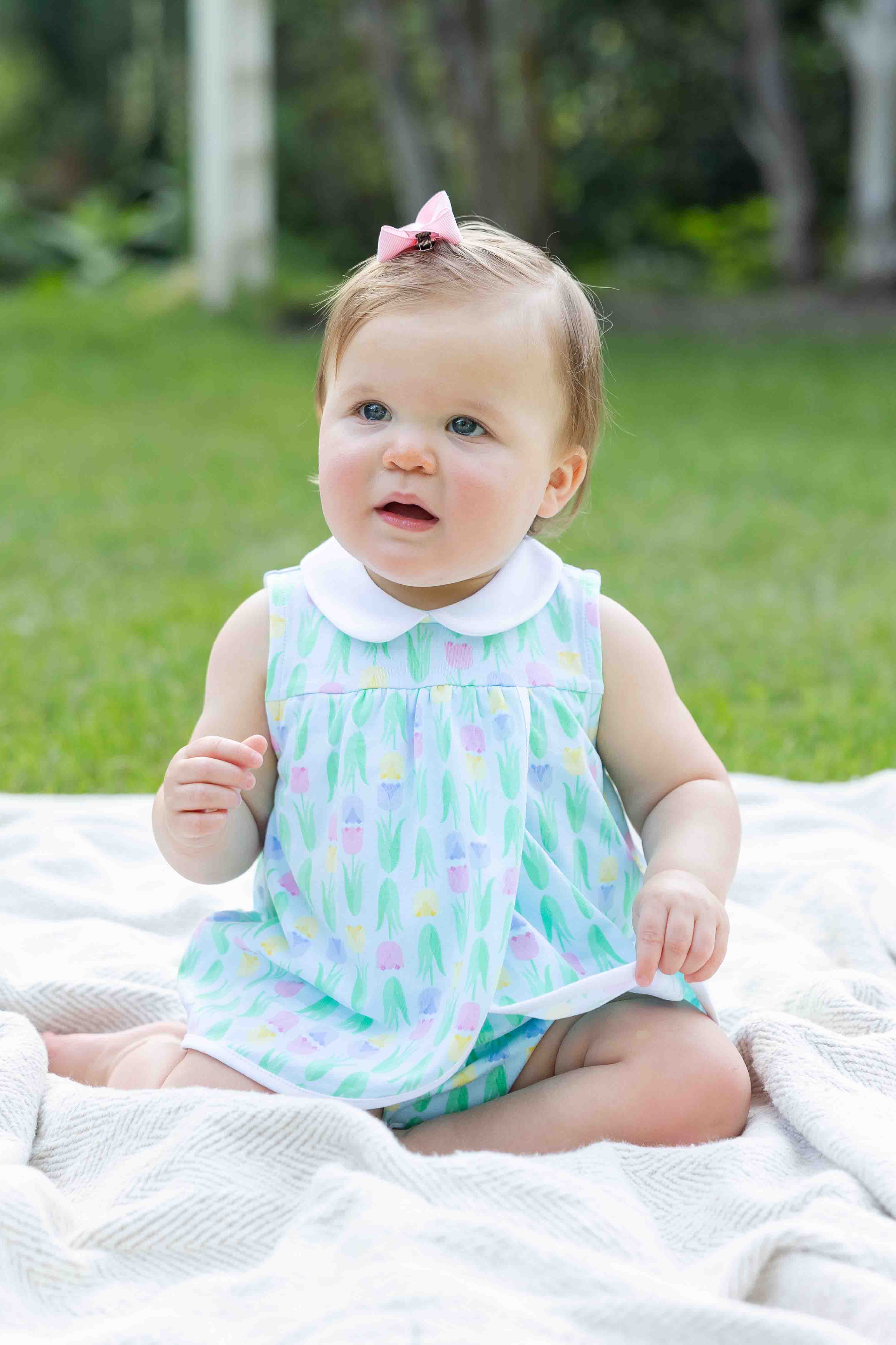 Baby sitting on a blanket outdoors wearing a colorful dress.