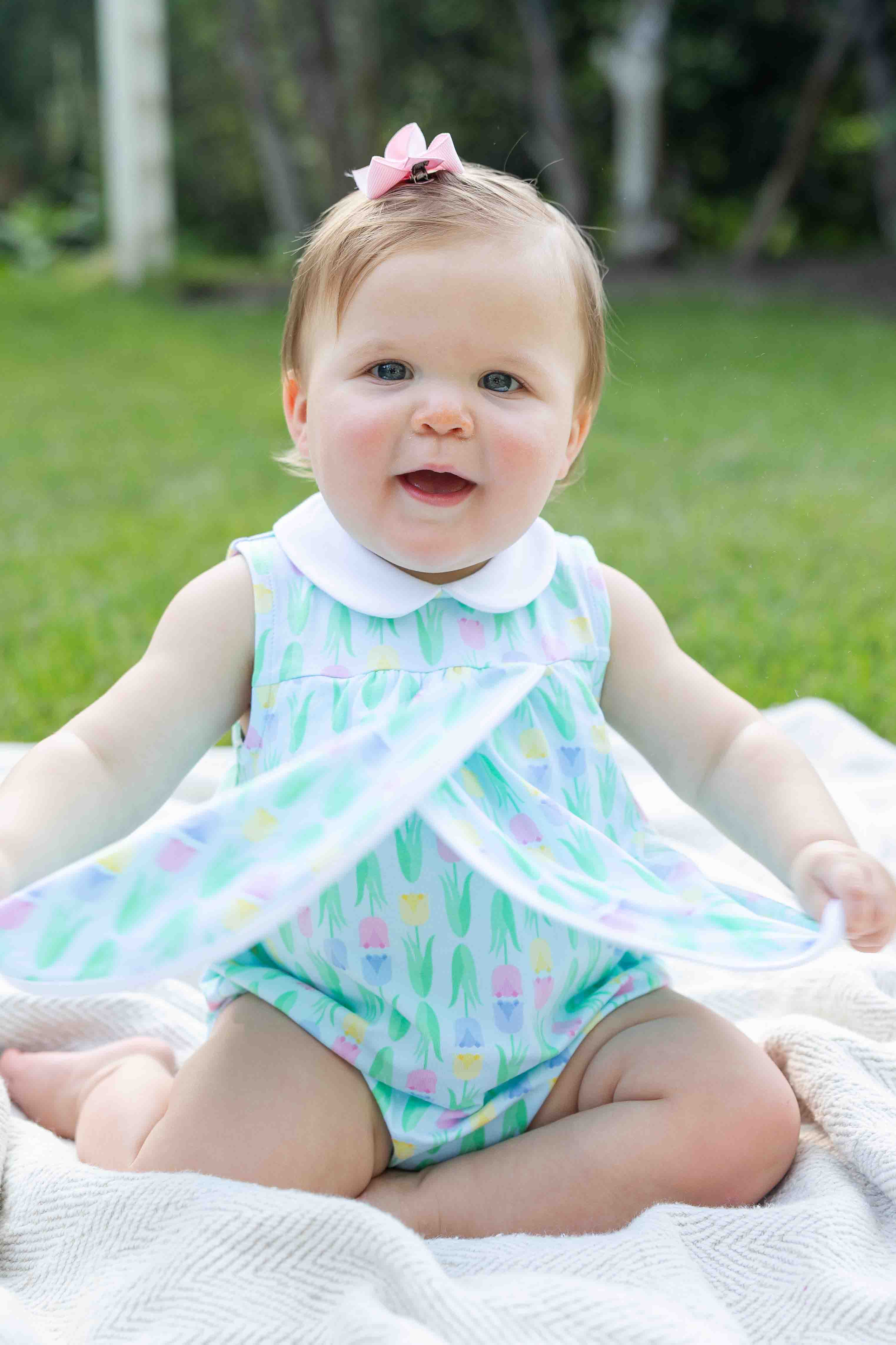 Baby sitting on a blanket outdoors wearing a colorful dress.