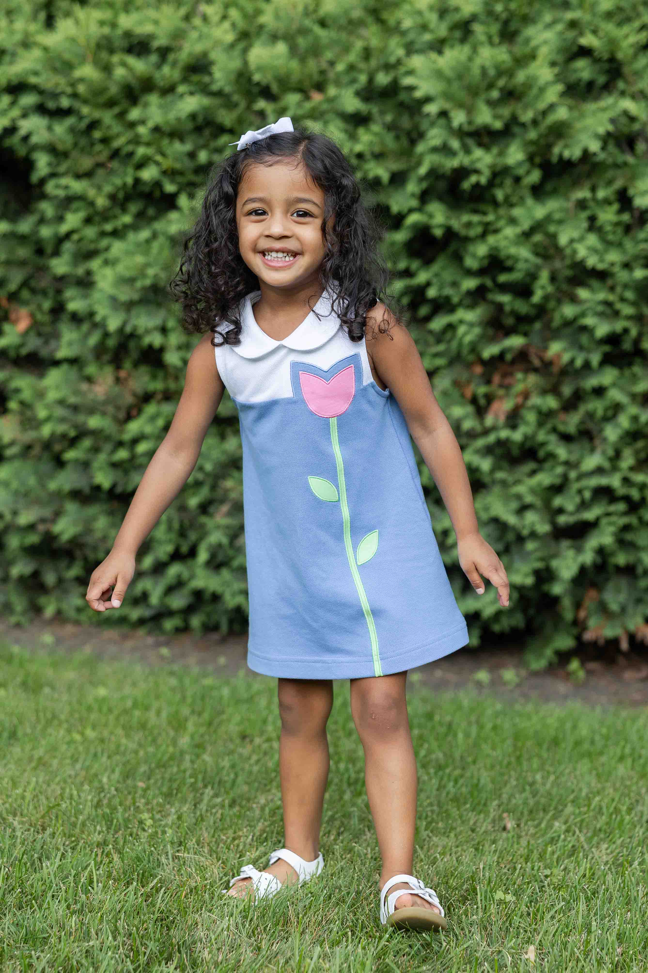 Young girl in a blue dress with a pink flower design standing on grass with green bushes in the background