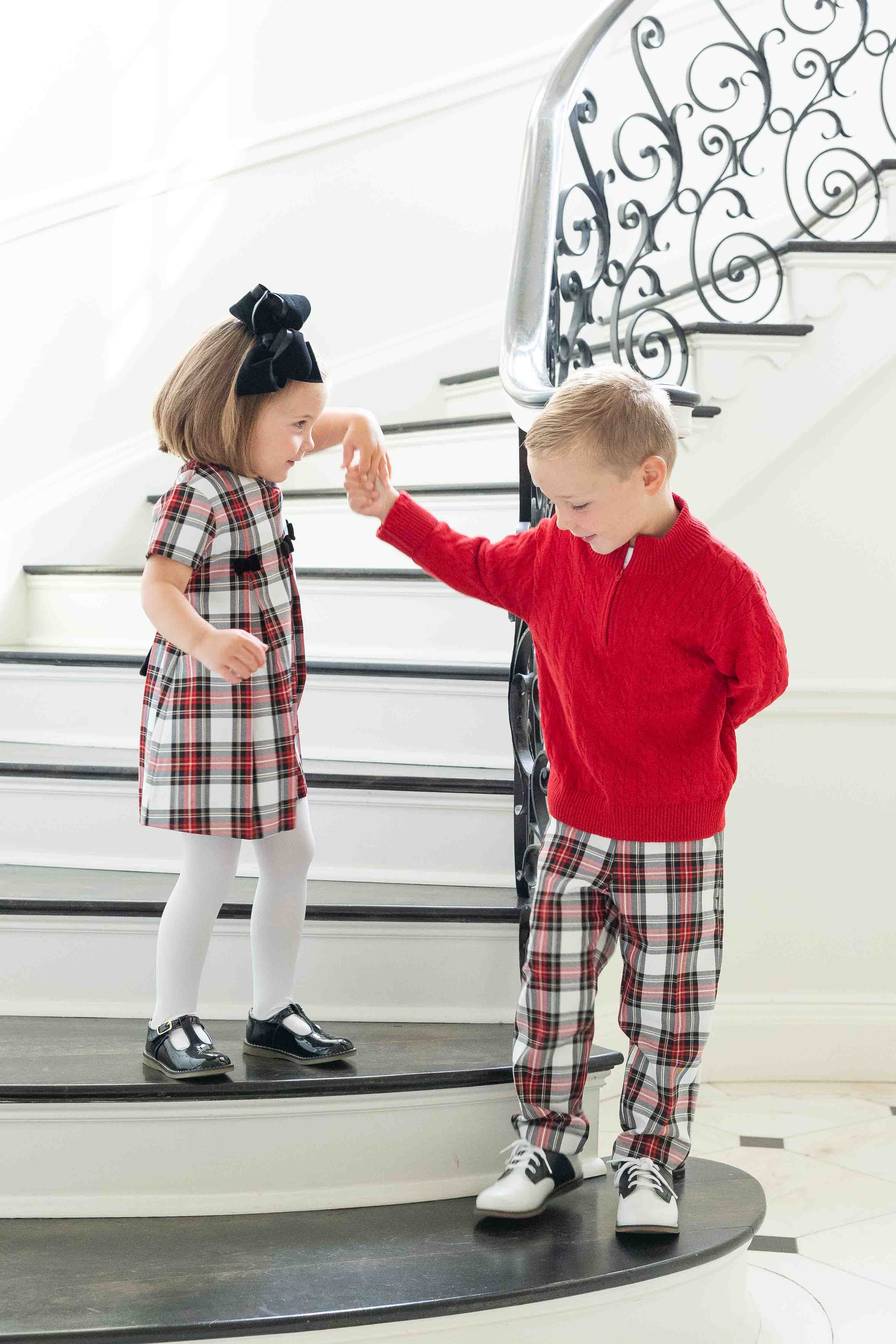 Two children, a girl in a plaid dress and a boy in a red sweater and plaid pants, standing on a staircase.