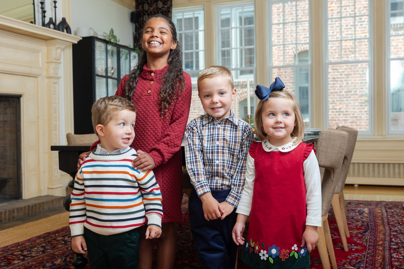Four children standing together in a room with a fireplace and large window.