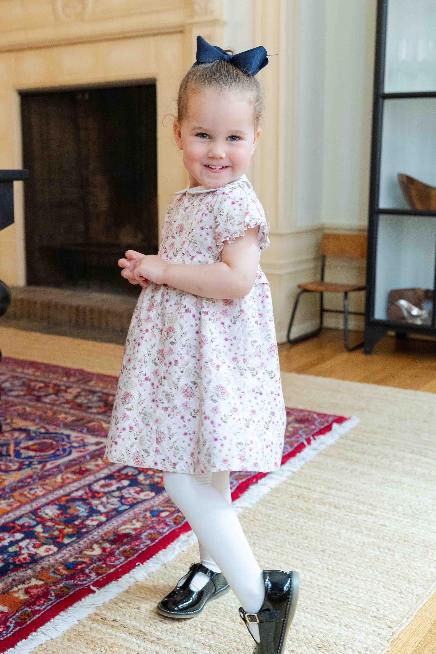 Young girl in a floral dress with a bow standing in a room with a fireplace and rug.