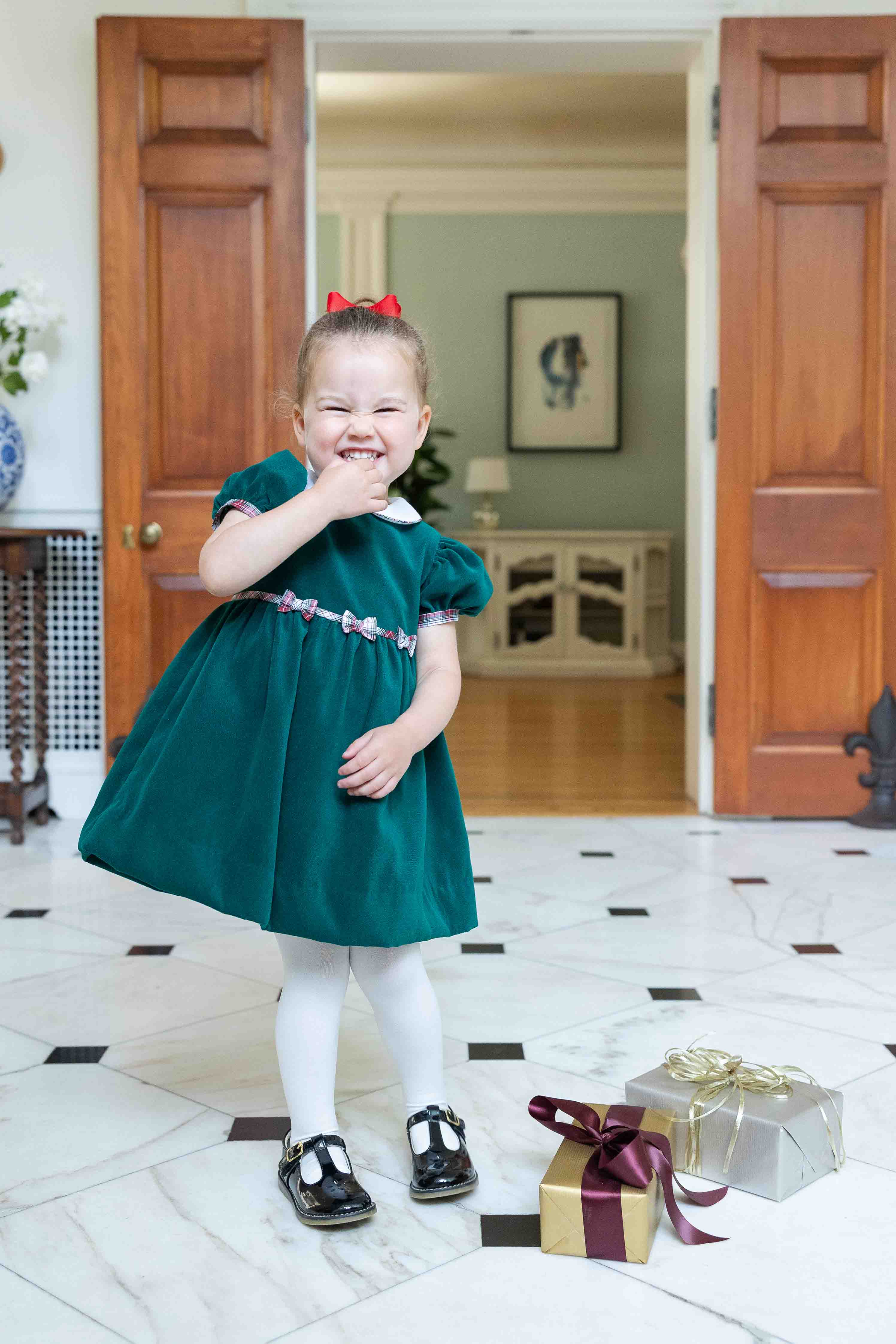 Child in a green dress standing in a room with wooden doors and a gift box.