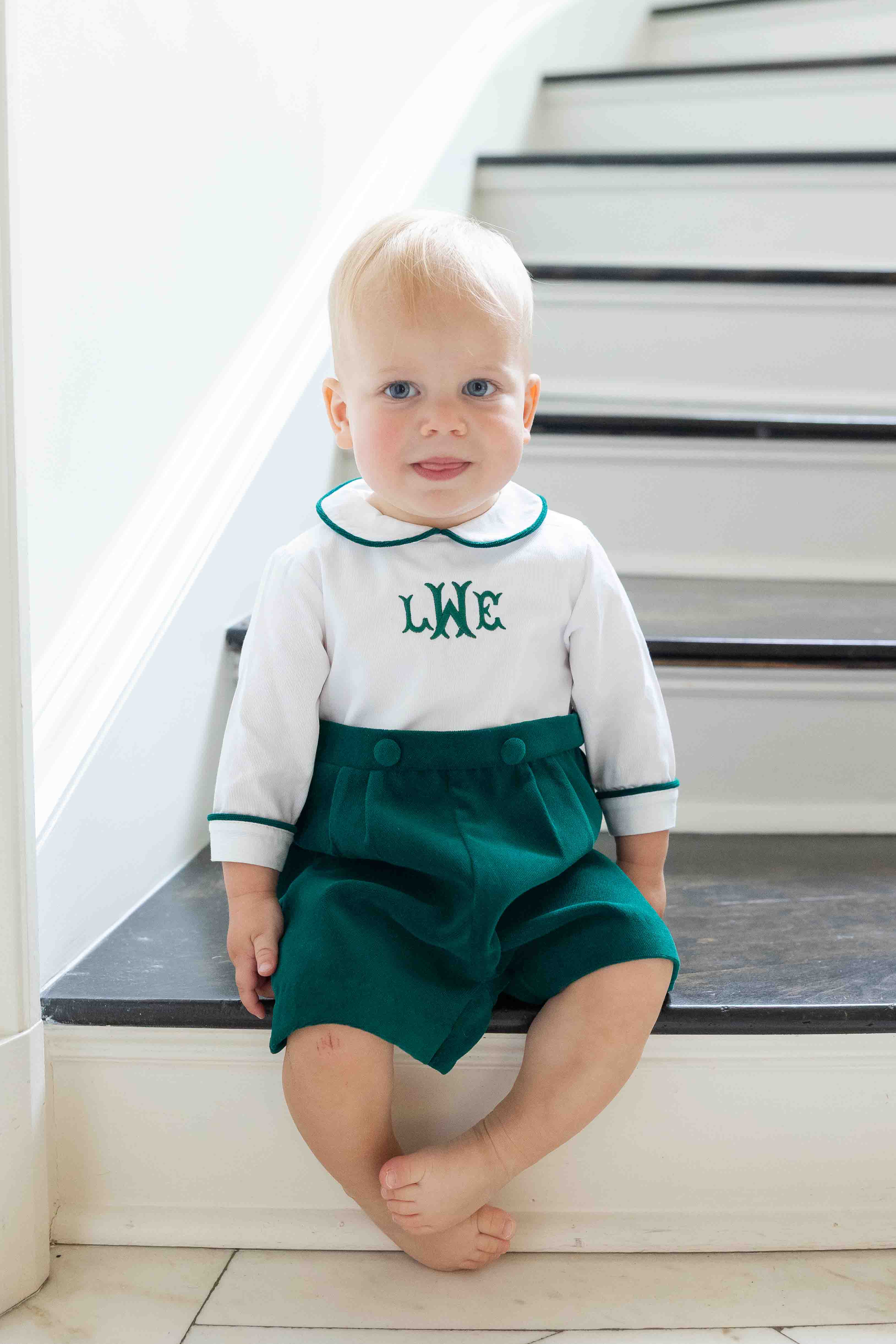 Baby sitting on a staircase wearing a shortall with a white top with green monogram and green shorts.