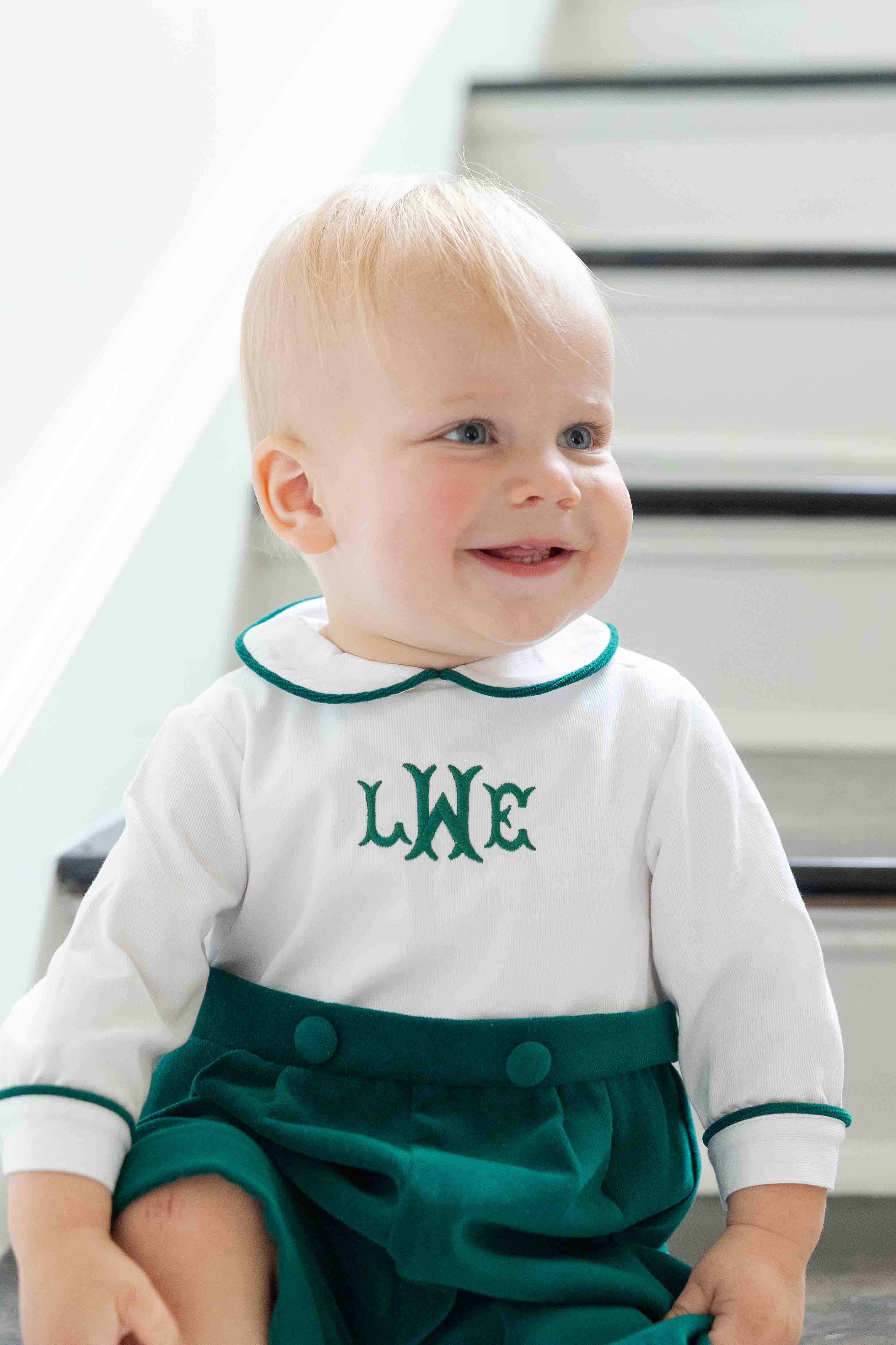 Baby wearing a shortall with a white top with green trim and green bottom, sitting on stairs.