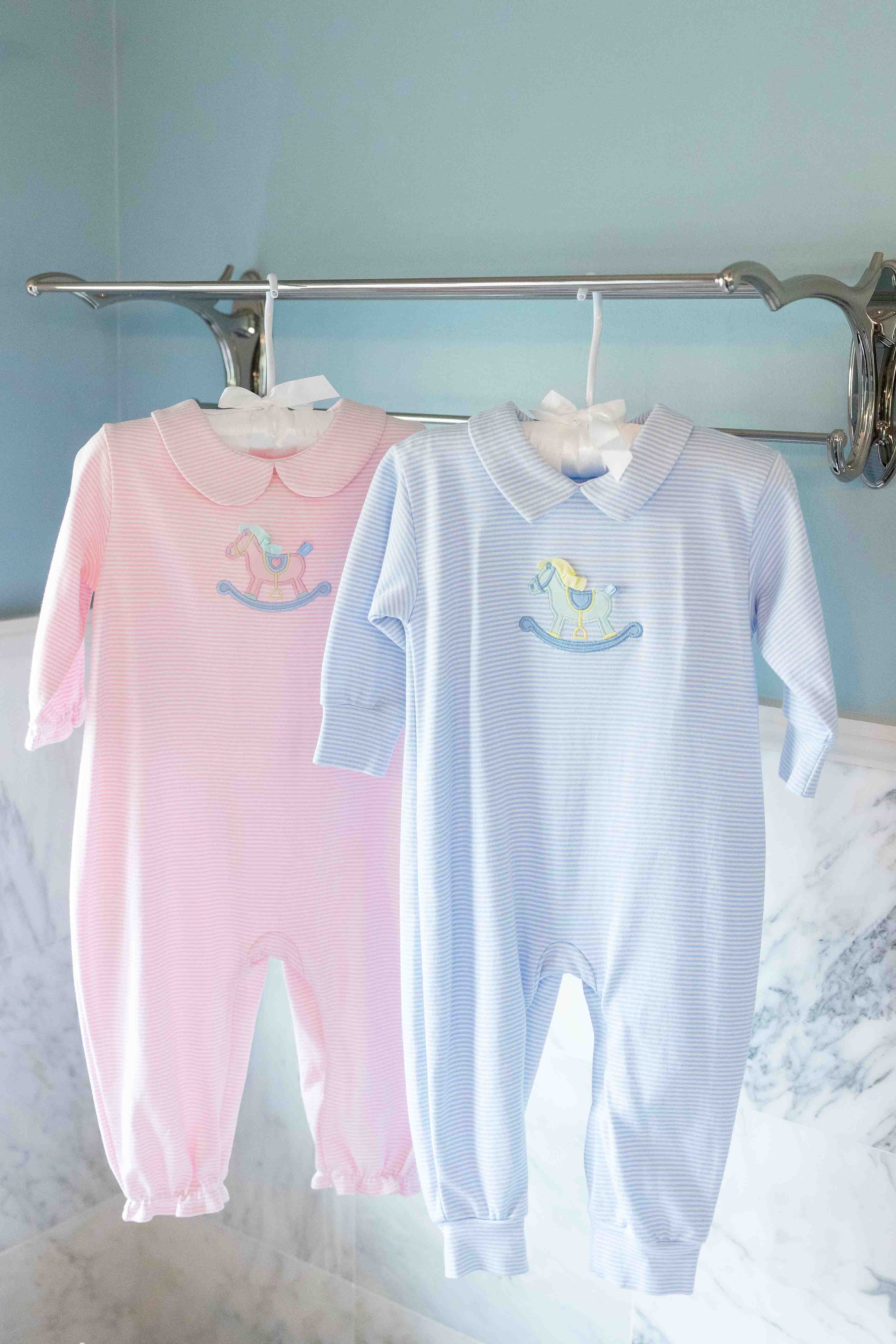 Pink and blue baby rompers hanging on a rack against a light blue wall.
