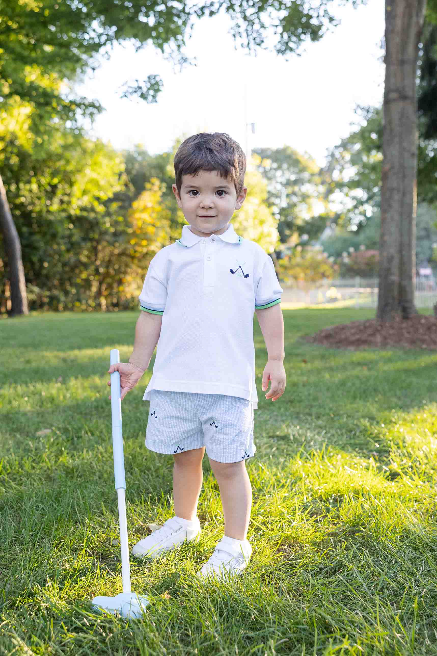 Young boy in a white polo shirt and light shorts standing on grass holding a golf club.